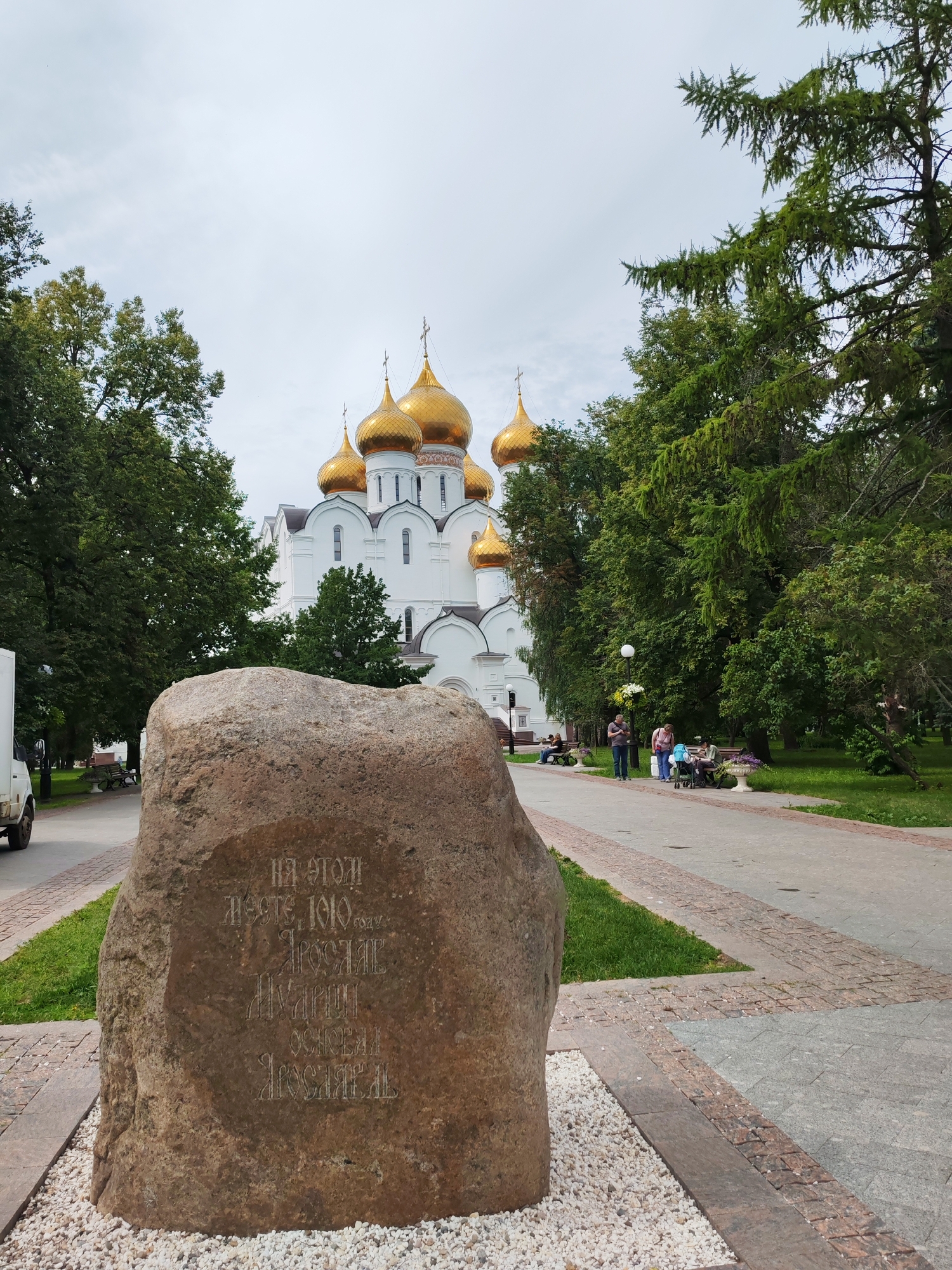 A large, rough-hewn stone with inscribed text stands in the foreground of a park area. The text on the stone, in Cyrillic script, appears to be historical and mentions dates and names. Behind the stone, a white building with golden domes, resembling a church, is visible, surrounded by lush green trees. Several people are seated on benches and walking along a paved path in the middle ground. The sky is overcast. The scene depicts an outdoor public space with architectural and natural elements, likely a historical or religious site in a park.