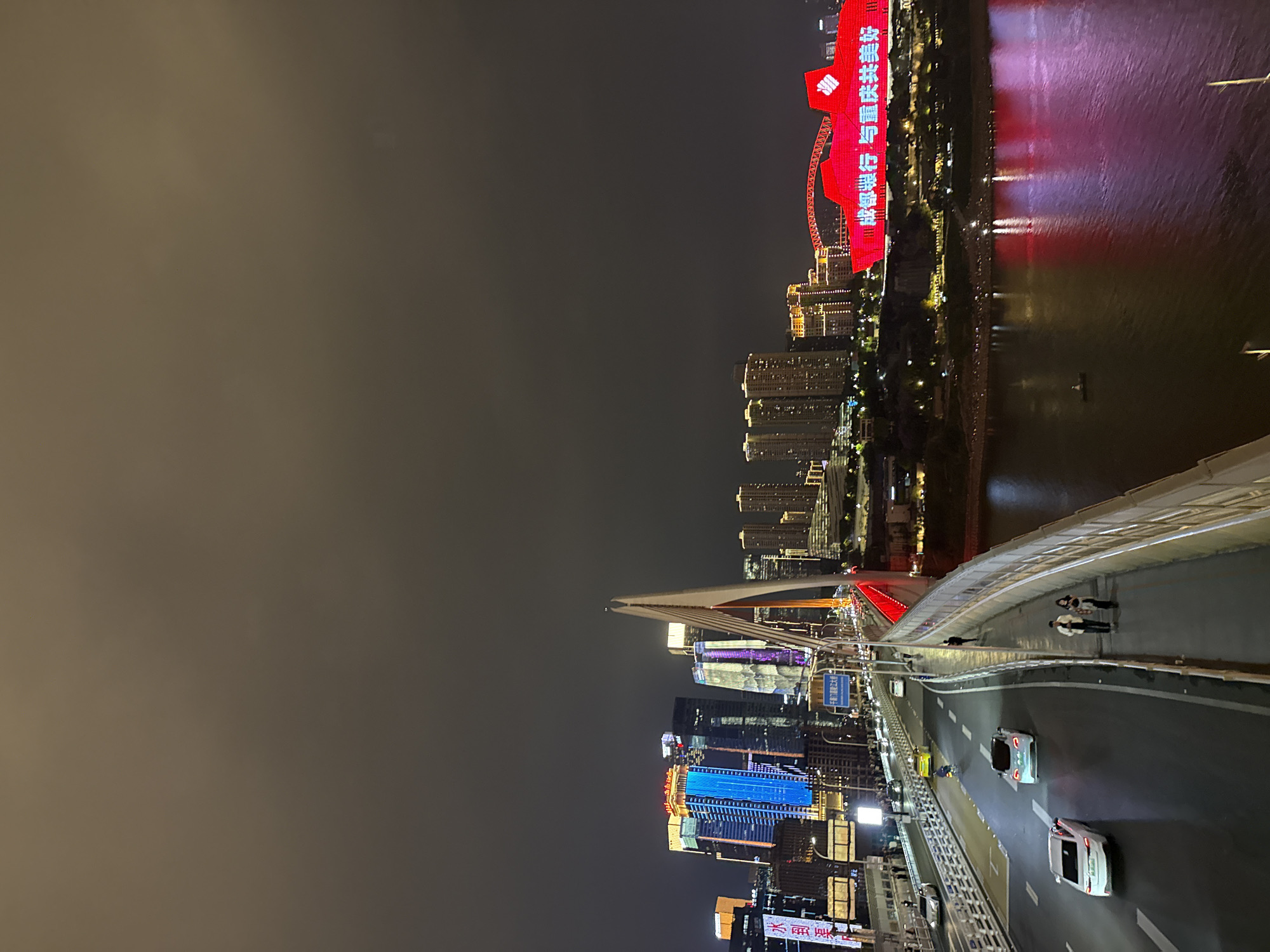 Night view from the Qiansimen Bridge in Yuzhong District, Chongqing, China. Several vehicles travel across the bridge. People are walking on the sidewalk, located on the bridge. In the distance, there are illuminated buildings and the river reflecting the light.