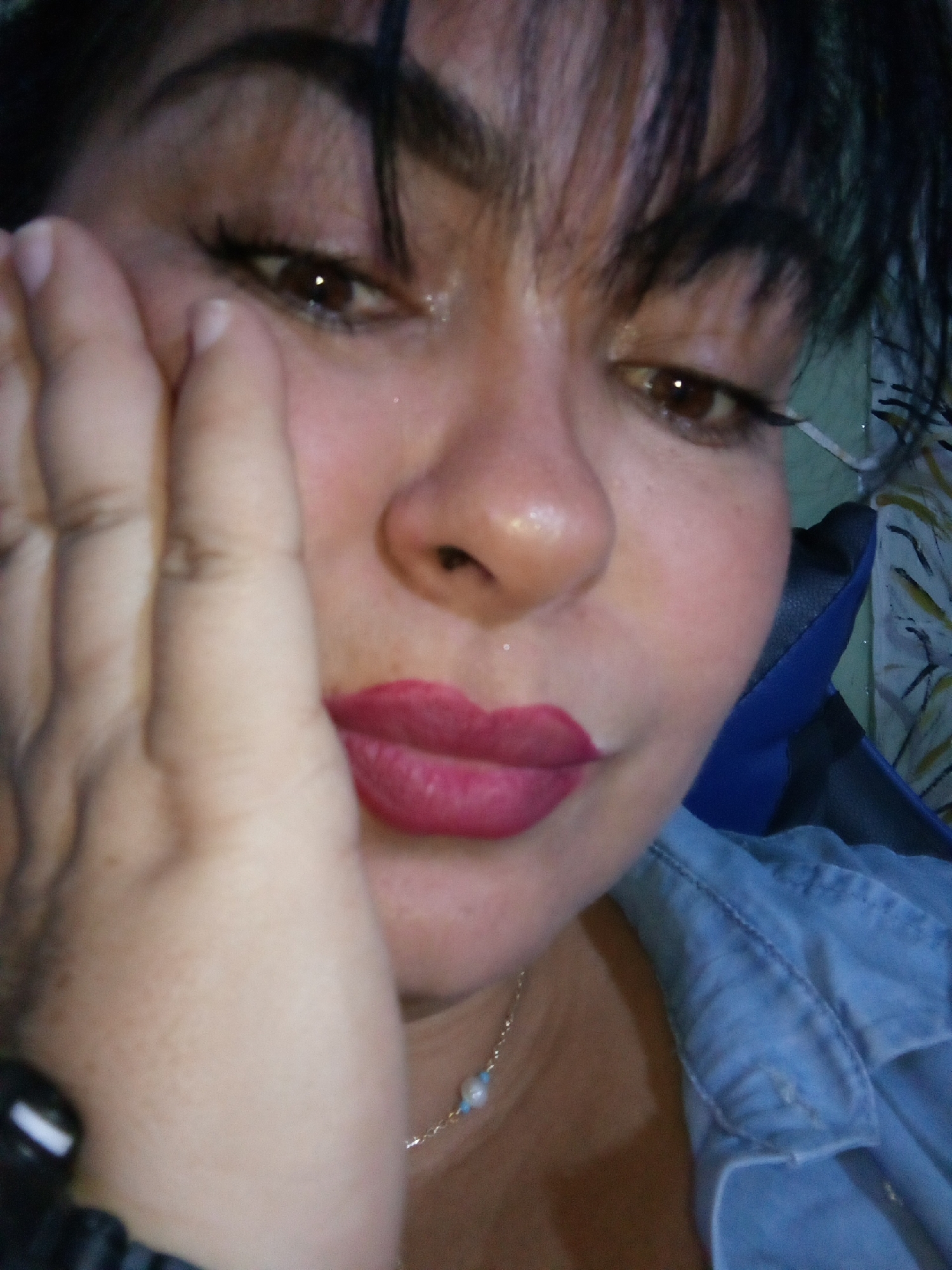 A close-up, slightly angled portrait features a woman with dark hair and brown eyes, possibly taken in a casual indoor setting in Juan Griego, Venezuela. Her head is tilted slightly, and her gaze is directed downwards and to her right, giving her expression a pensive or contemplative quality.
She has dark, well-groomed eyebrows and her eyes are accentuated with eyeliner and mascara. Her lips are covered in a vibrant red or fuchsia lipstick. Her right hand rests against the left side of her face, with fingers partially obscuring her cheek, creating a relaxed pose.
She is wearing a light blue, possibly denim, collared shirt. Around her neck, a delicate silver-colored chain necklace is visible, adorned with small light blue beads and a single white pearl-like bead. A part of a black band, possibly a watch or bracelet, is seen on her left wrist in the bottom left corner of the frame.
The background is softly blurred, showing indistinct patterns and colors, including a segment of a blue object, possibly part of a chair or headrest, behind her. There are no strong cues for the time of day or specific weather conditions. No visible text is present in the image.