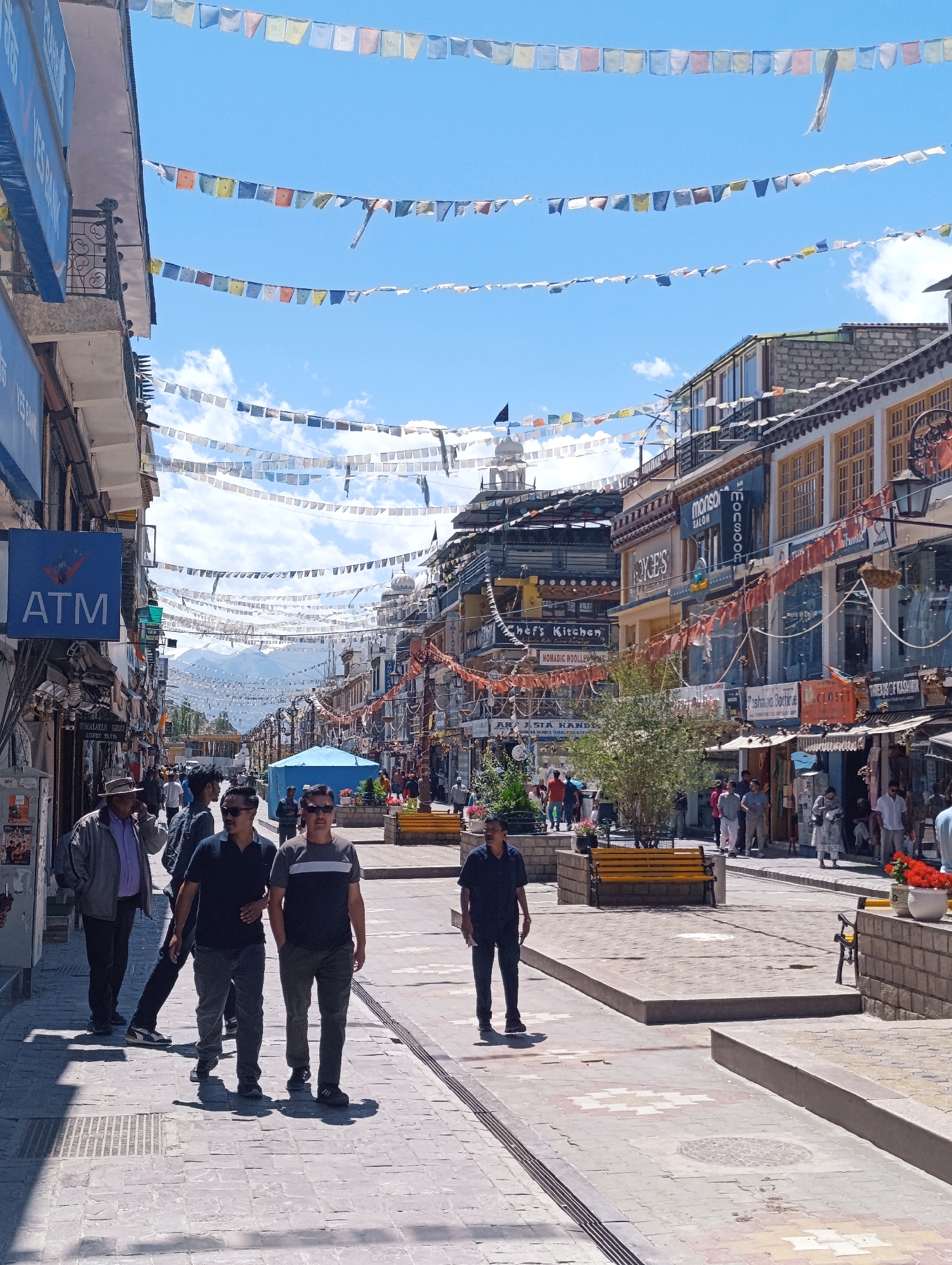The image depicts a street scene in Leh, India, characterized by colorful prayer flags strung across the sky between buildings. The street is paved with stone tiles and lined with shops, some of which have awnings and display windows. A blue ATM sign is visible on the left side of the street. Several people are walking along the street, some individually and others in small groups. In the background, mountains are visible under a partly cloudy sky. The architecture suggests a town or city setting with a mix of traditional and modern elements.
