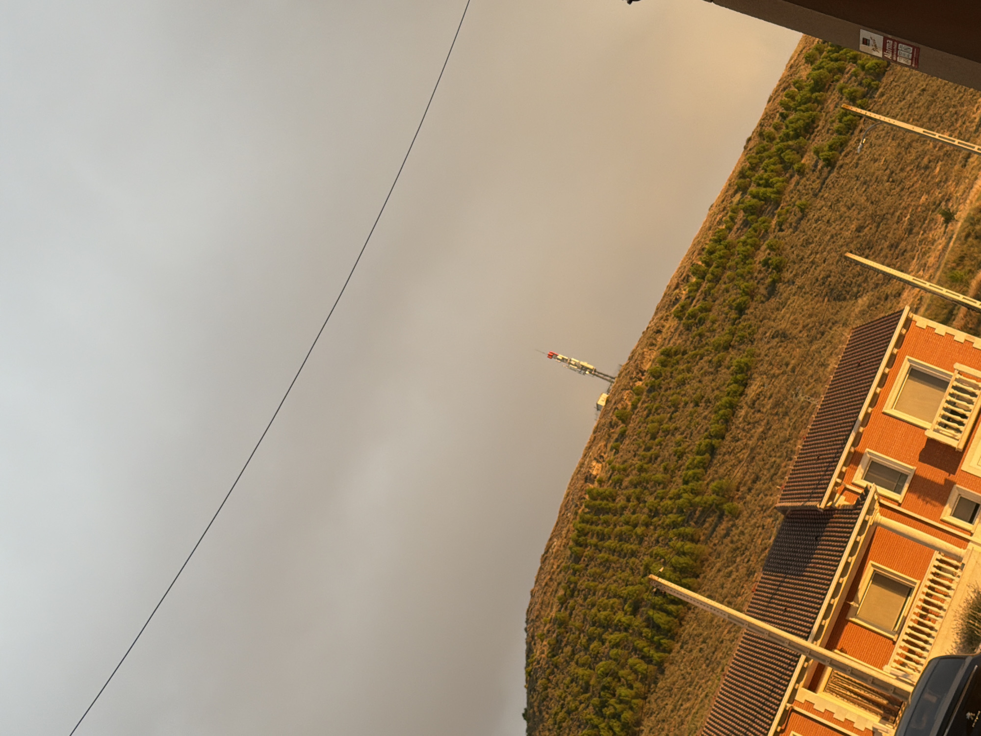 The image, taken in Vallelado, Spain, captures a hillside covered in dense, green trees, receding into the distance under a hazy, golden sky. The hillside is intersected by a series of wooden poles, some of which are visible in the foreground. A communication tower with red and white sections stands prominently on the crest of the hill. In the lower left of the frame, a portion of an orange brick building with a tiled roof is visible. The building features several windows with white railings and a balcony. A black car is partially visible in the bottom left corner. The overall scene is characterized by warm, diffused lighting, suggesting it was taken during sunset or sunrise. A thin, dark line, possibly a power line or cable, diagonally bisects the sky.