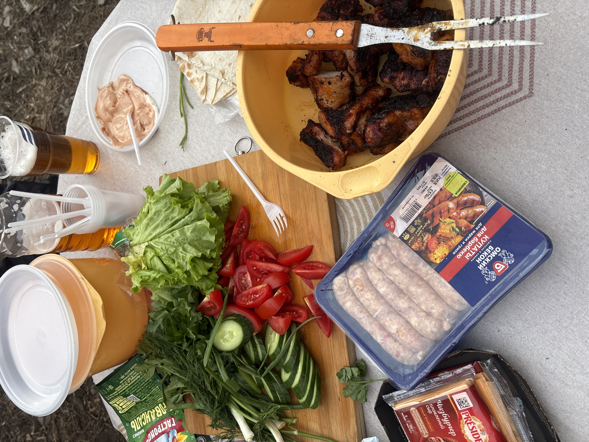 The image shows a table laden with food, likely set for an outdoor meal or picnic. A yellow bowl contains grilled meat, accompanied by a large orange handled carving fork. A wooden cutting board holds an assortment of fresh vegetables including sliced tomatoes, cucumbers, and green onions, alongside leafy lettuce and various herbs. Next to the board is a packaged container of raw sausages, with the product name visible as "Omsky Bacon Kupaty" in Russian, intended for grilling. Also present is packaged processed cheese, specifically "Président Cheeseburger" slices. Cups and plastic utensils suggest a casual, disposable setup. Beverages are present in plastic cups, with one appearing to contain beer. A container of sauce is also visible. The overall setting seems to be informal, likely outdoors, but there is no evidence to specifically determine that this scene takes place in Krasnoyarsk.