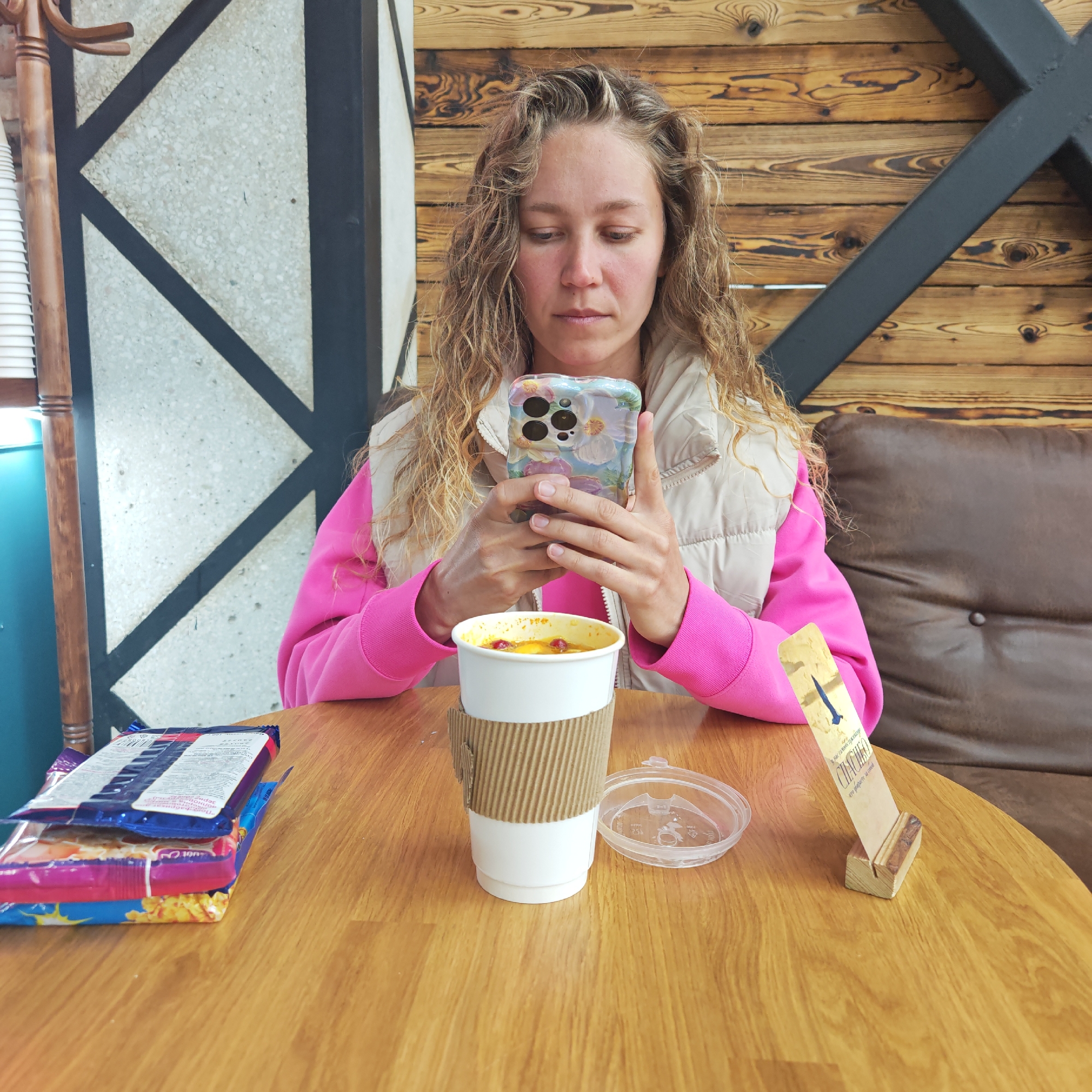 The photograph captures a woman seated at a round, wooden table, likely in a café in an unidentified city. She is light-skinned with long, curly blonde hair and is dressed in a pink sweatshirt under a light-colored vest. The woman is focused on a smartphone held in her hands. On the table, there is a stack of packaged food items, a white paper cup with a corrugated cardboard sleeve, and a small wooden stand holding a card with text printed on it. The background includes a wall with a geometric design and wooden paneling. A portion of a brown leather couch is visible to the right of the woman.