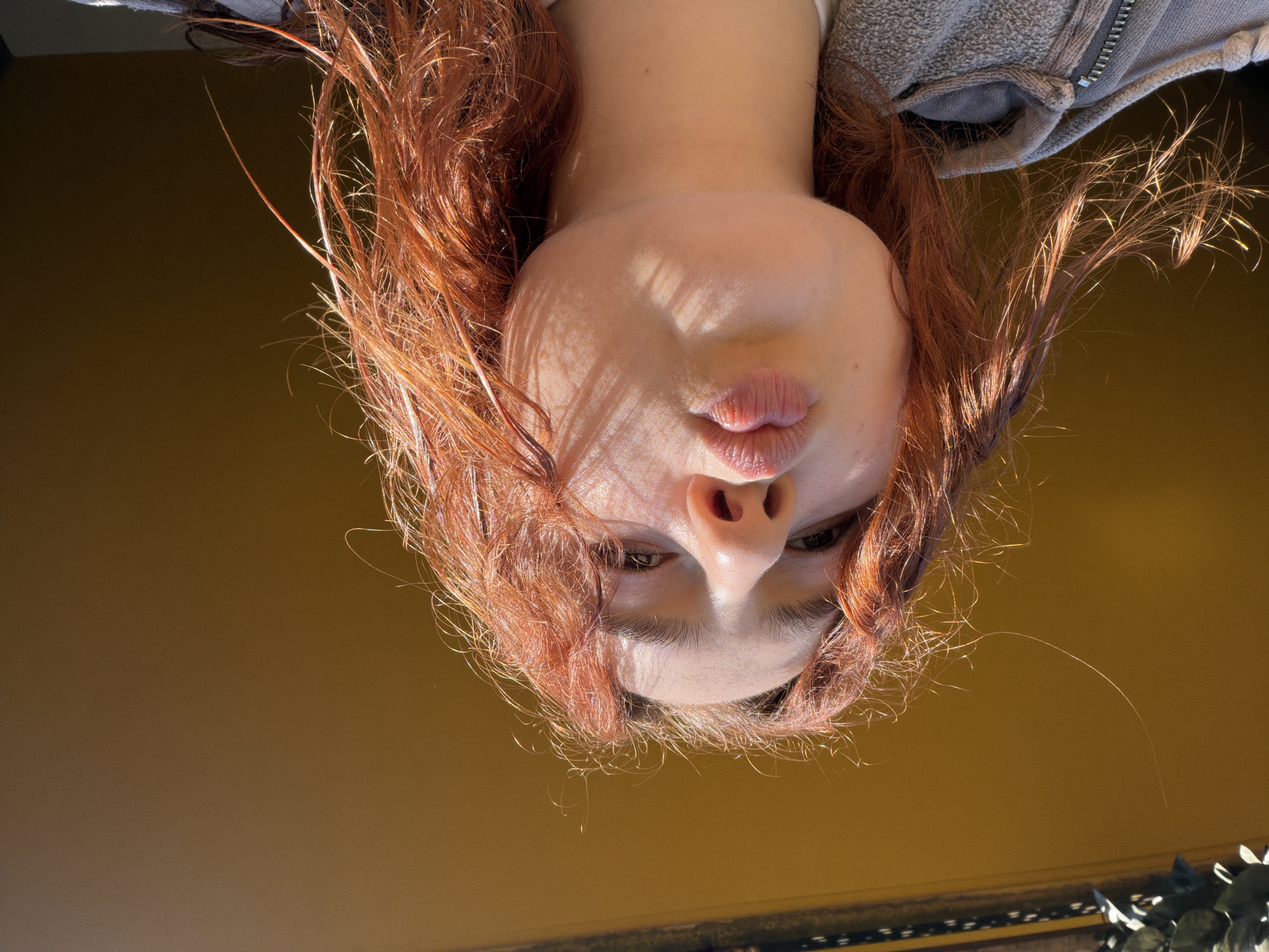 The image is an upside-down, close-up view of a person's face, likely a young woman with auburn hair. Her face is tilted forward and towards the camera, revealing her nose, lips in a pouting expression, and partially visible eyes. Sunlight and shadows are cast across her face. The background appears to be a plain, possibly yellowish surface. A portion of the individual's gray zippered hoodie is also in frame, with hints of a metallic texture near the upper edge. There's no direct evidence of the location being in Strasbourg, France. Further context would be needed to confirm that aspect of the description.