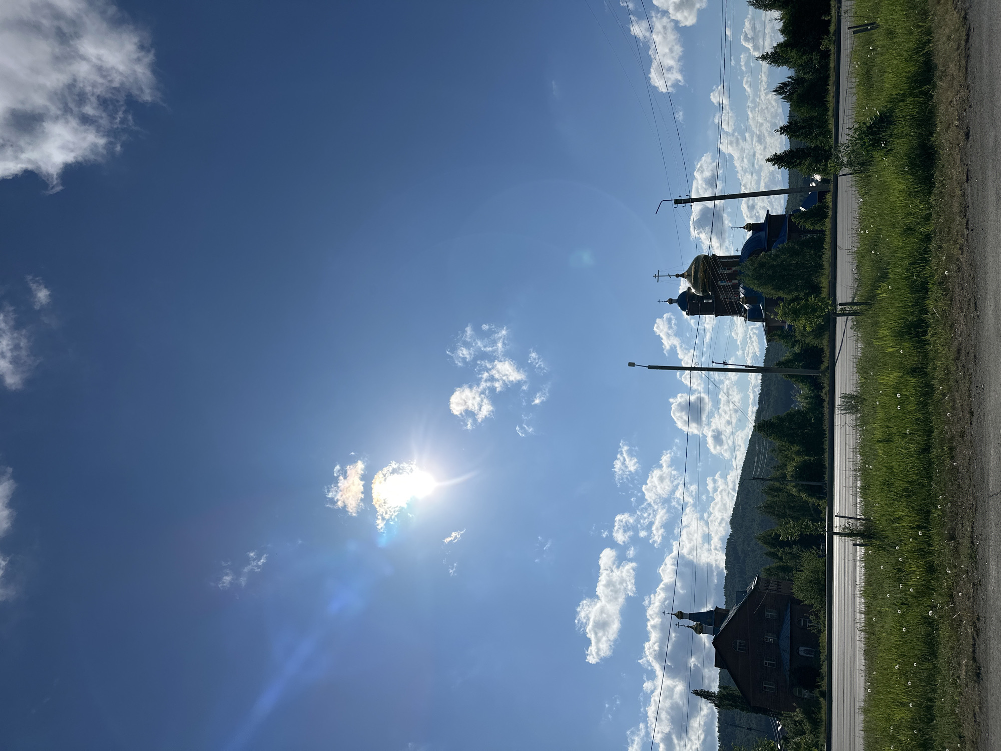 The image shows a sunny, daytime scene with a blue sky filled with cumulus clouds. The sun is visible, creating a bright glare. In the distance, there is a church with golden domes and crosses, and another building with a steeple. There are trees and grass along the side of a road. Power lines stretch across the sky. The location is in an unknown city.