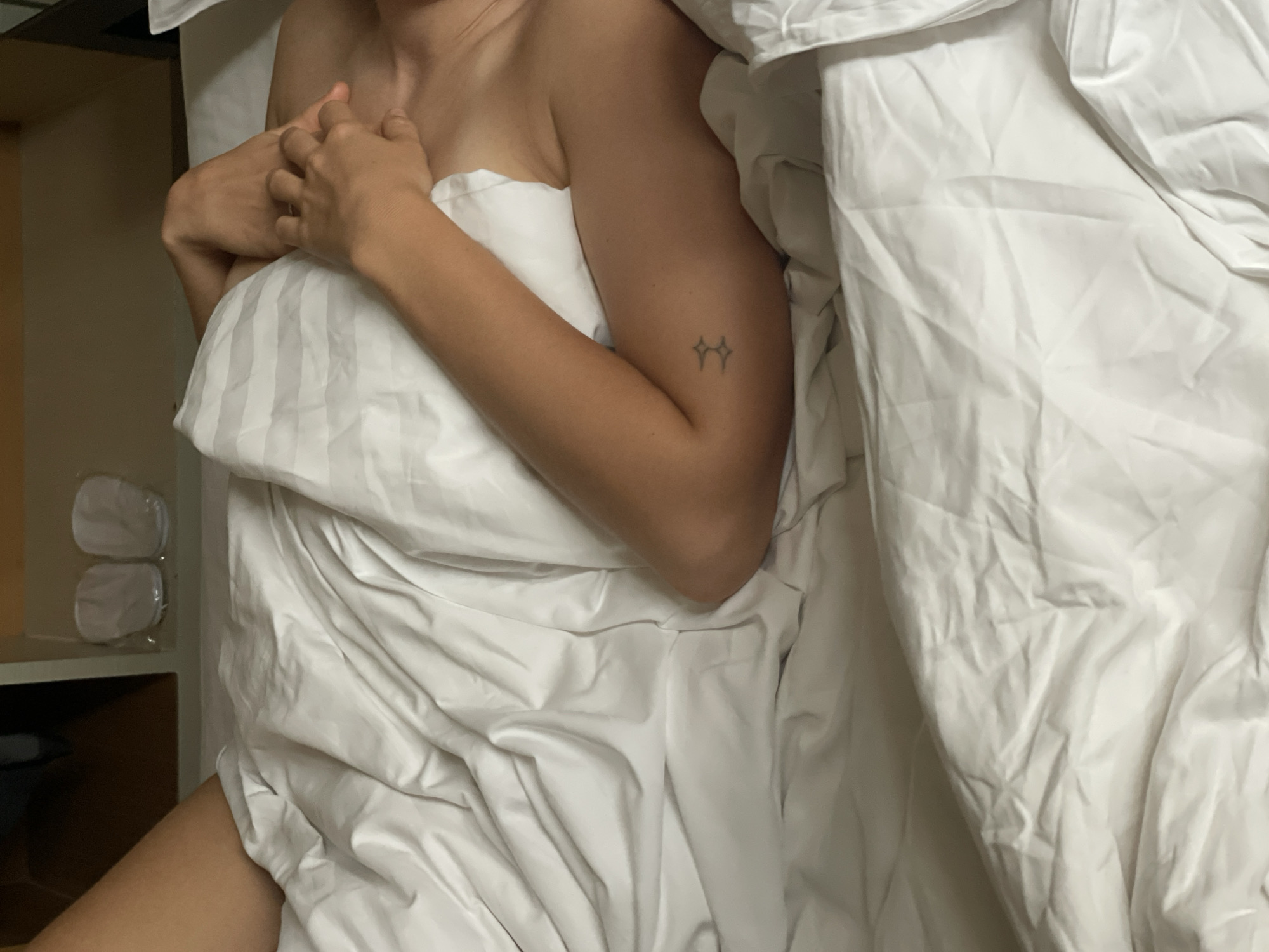 An overhead shot captures a person resting in bed, partially covered by white bedding, likely within a hotel room in Bangkok, Thailand. The subject, implied to be female, is positioned centrally, with their bare right shoulder and arm visible. Their hands are clasped gently near their chest. A small, stylized tattoo of two linked star-like symbols is visible on their right arm.
The bed is adorned with white sheets and a white comforter or duvet, some sections of which feature subtle striped patterns. A portion of the person's bare thigh is visible near the bottom left corner of the frame. To the left of the bed, on a shelf or bedside table, two pairs of white slippers in clear plastic packaging are visible, reinforcing the hotel setting. The lighting is soft, contributing to a calm and relaxed atmosphere. No explicit text is visible beyond the tattoo symbol.