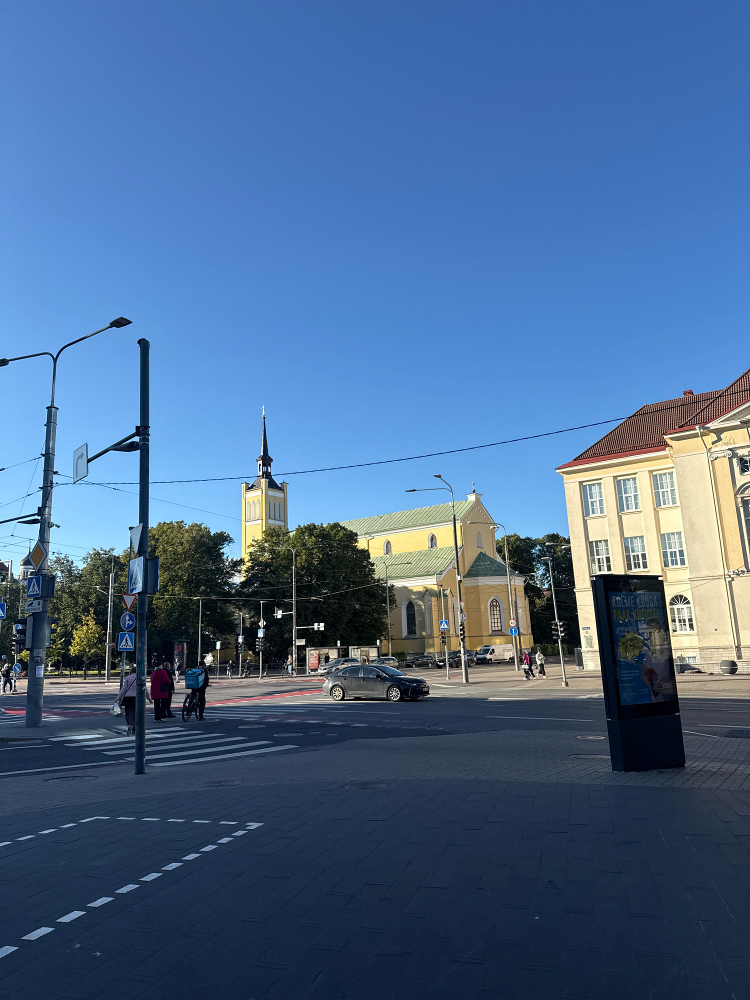 This is a daytime, eye-level, outdoor shot of a city street in Tallinn, Estonia. The scene is dominated by a clear, bright blue sky. In the mid-ground, a large, yellow church with a green roof and a tall, black steeple stands prominently. To the right of the church is a pale yellow building with a red tiled roof and multiple windows. A black, digital advertising kiosk is positioned in the foreground to the right. The street features a crosswalk with white stripes and dark grey paving stones. Several people are visible, including a person riding a bicycle with a large blue delivery bag on their back, and a few pedestrians crossing the street. Streetlights and traffic signals are present, along with various street signs. The overall environment appears to be a busy urban intersection on a sunny day.