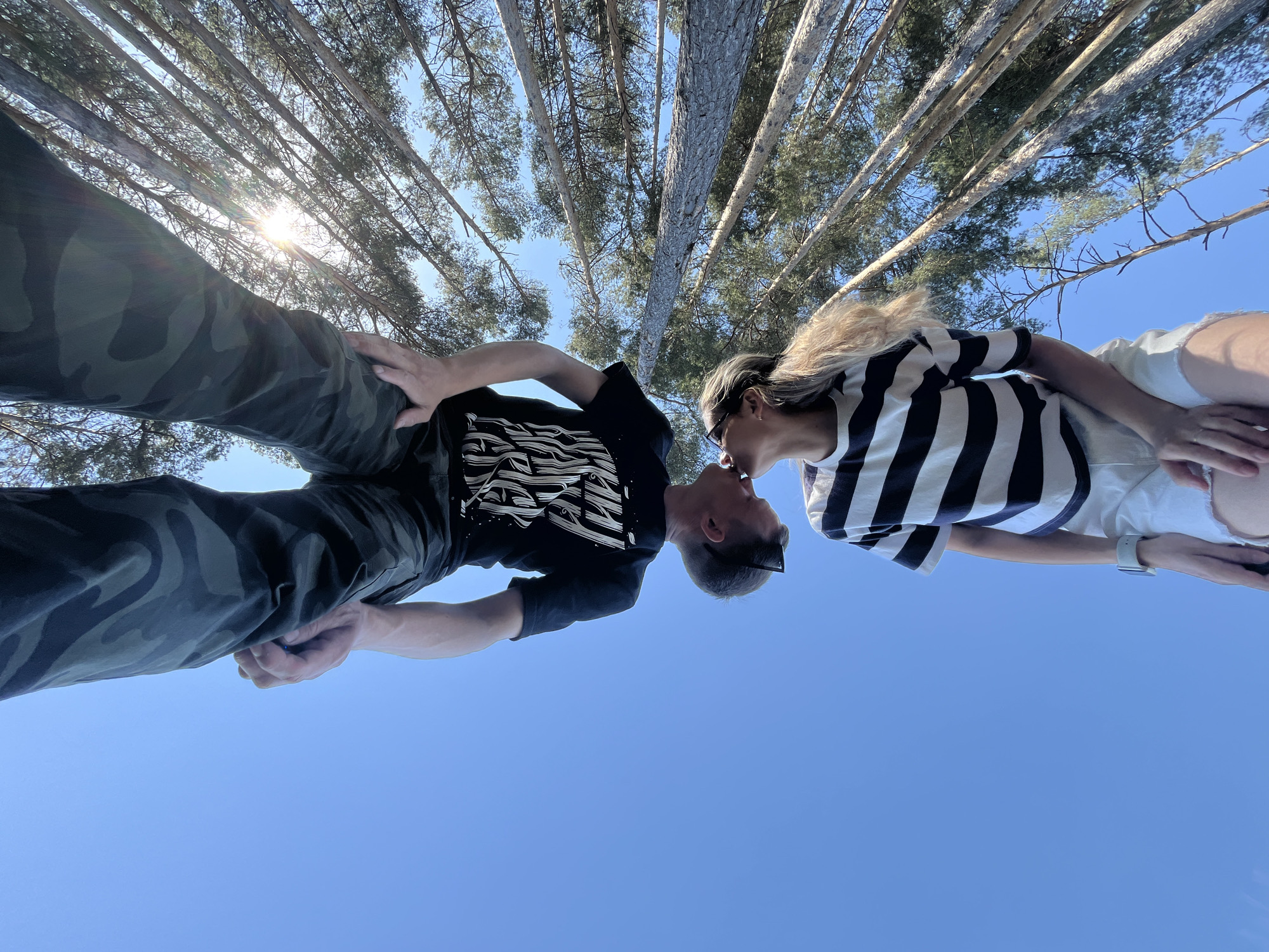 A couple is kissing outdoors, framed by a low-angle shot that includes tall, thin trees reaching upwards towards a clear, bright blue sky. Sunlight is visible through the trees. The person on the left wears camouflage pants and a black t-shirt with a white graphic design. The person on the right wears a striped black and white shirt and white shorts. The location is Chaikovsky, Russia.