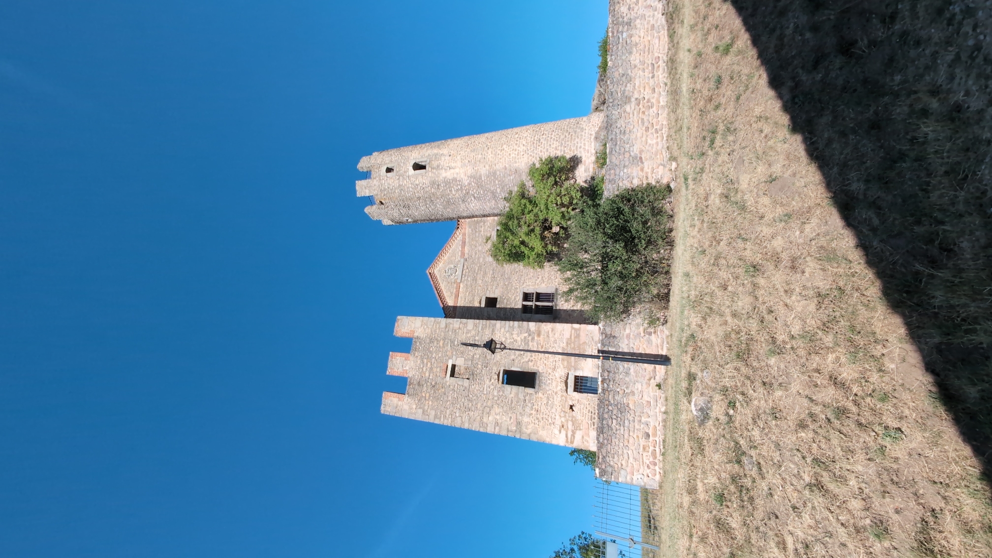 The image captures a low-angle view of the Château de Chambles, a medieval castle situated on a hill in Chambles, France. The castle is constructed of stone and features two prominent towers, one round and one rectangular, both with crenellations. The larger, rectangular tower on the left is angled slightly, while the round tower on the right leans significantly. Several windows are visible in the towers and the connecting building, some of which are dark or appear to be broken. A single black lamppost stands in front of the rectangular tower. Lush green trees and bushes are growing around the base of the castle. The castle is perched atop a grassy slope, with dry, yellow grass covering the foreground. A shadow cast by an unseen object falls across the lower right portion of the image. The sky above is a clear, vibrant blue, indicating a sunny day. A metal fence runs along the left side of the hill.