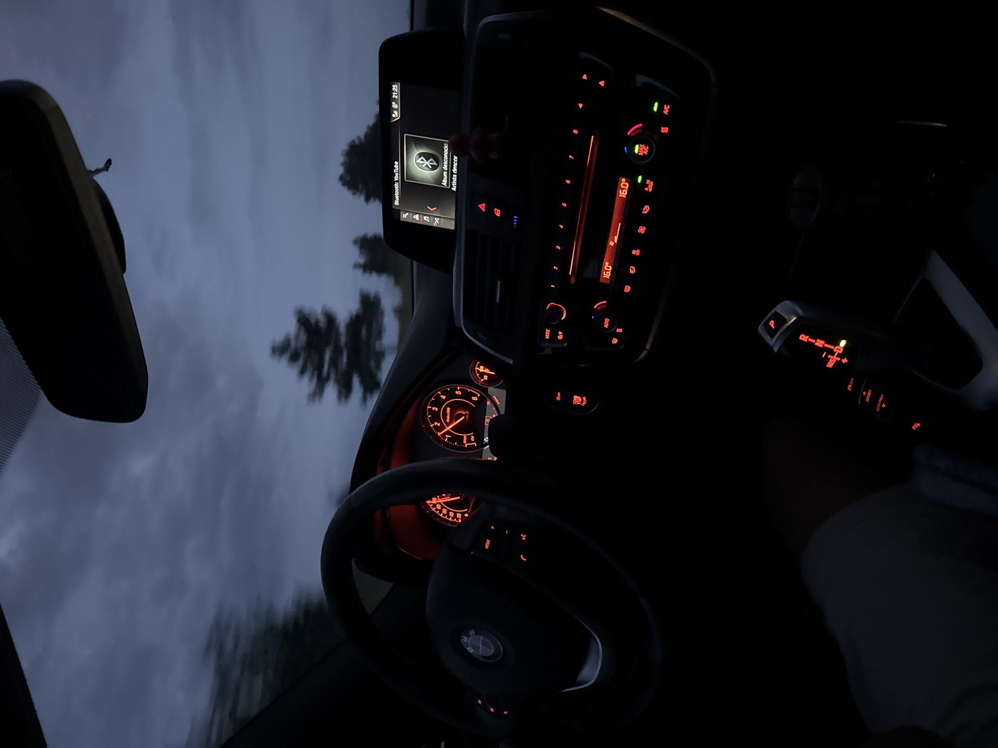 The image shows the interior of a car, likely at dusk or nighttime, with the dashboard and steering wheel illuminated by red and orange lights. The sky is visible through the windshield, with dark clouds and some trees silhouetted against it. The car's infotainment screen displays "Bluetooth: YouTube" and the time "21:25". The temperature is set to 16.0 degrees Celsius. A gear shifter with a "P" for Park is visible in the foreground. The scene appears to be in the city of Krepost, Bulgaria, based on the provided location. There are no people clearly visible in the image.