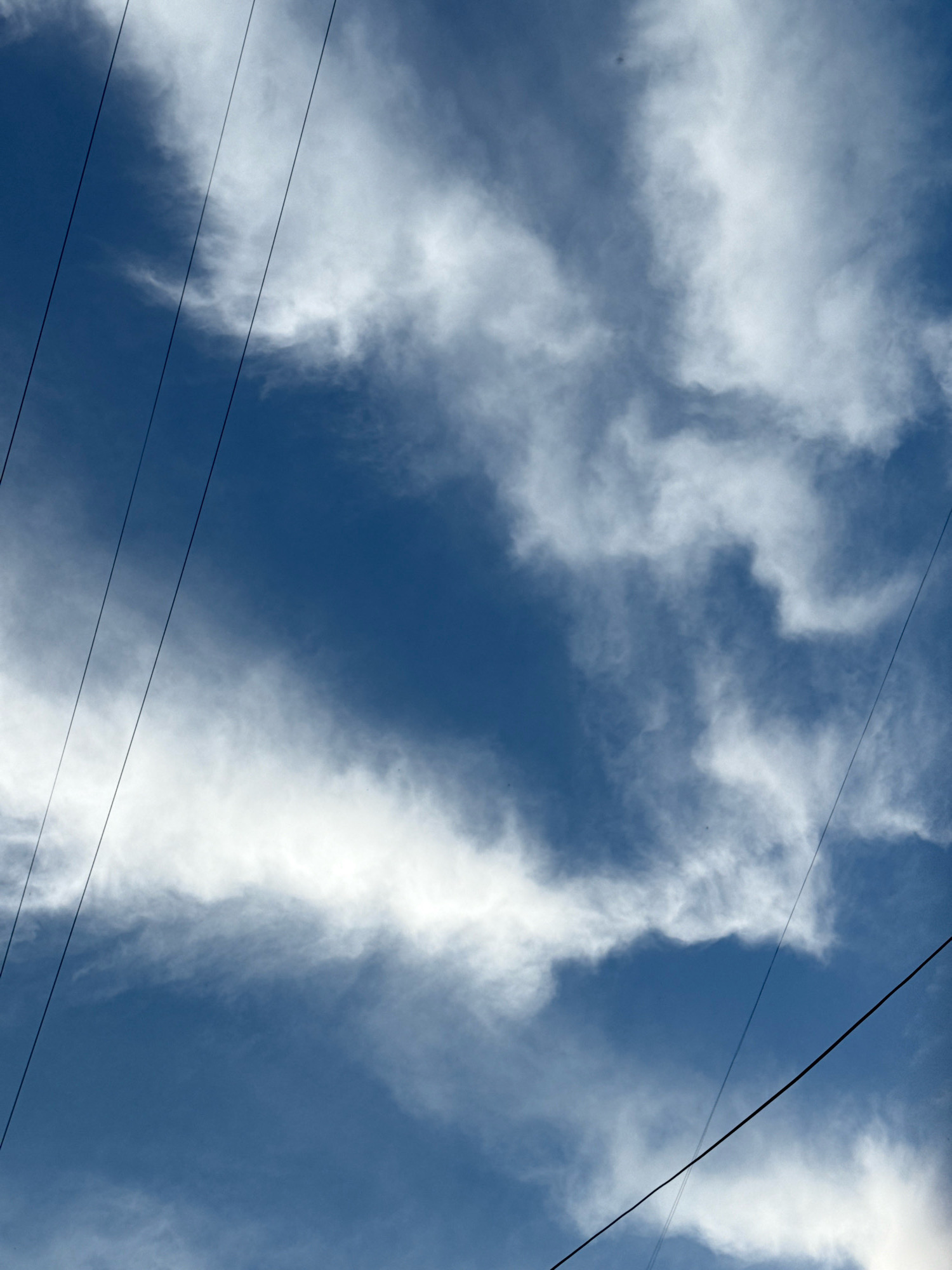 A view of the sky over Chaikovsky, Russia, featuring a deep blue expanse dotted with wispy white clouds. Several power lines stretch diagonally across the frame, obscuring portions of the sky and clouds. The clouds appear thin and scattered, with some areas exhibiting a more concentrated, streaky texture. The image is captured from a low angle, looking upwards.