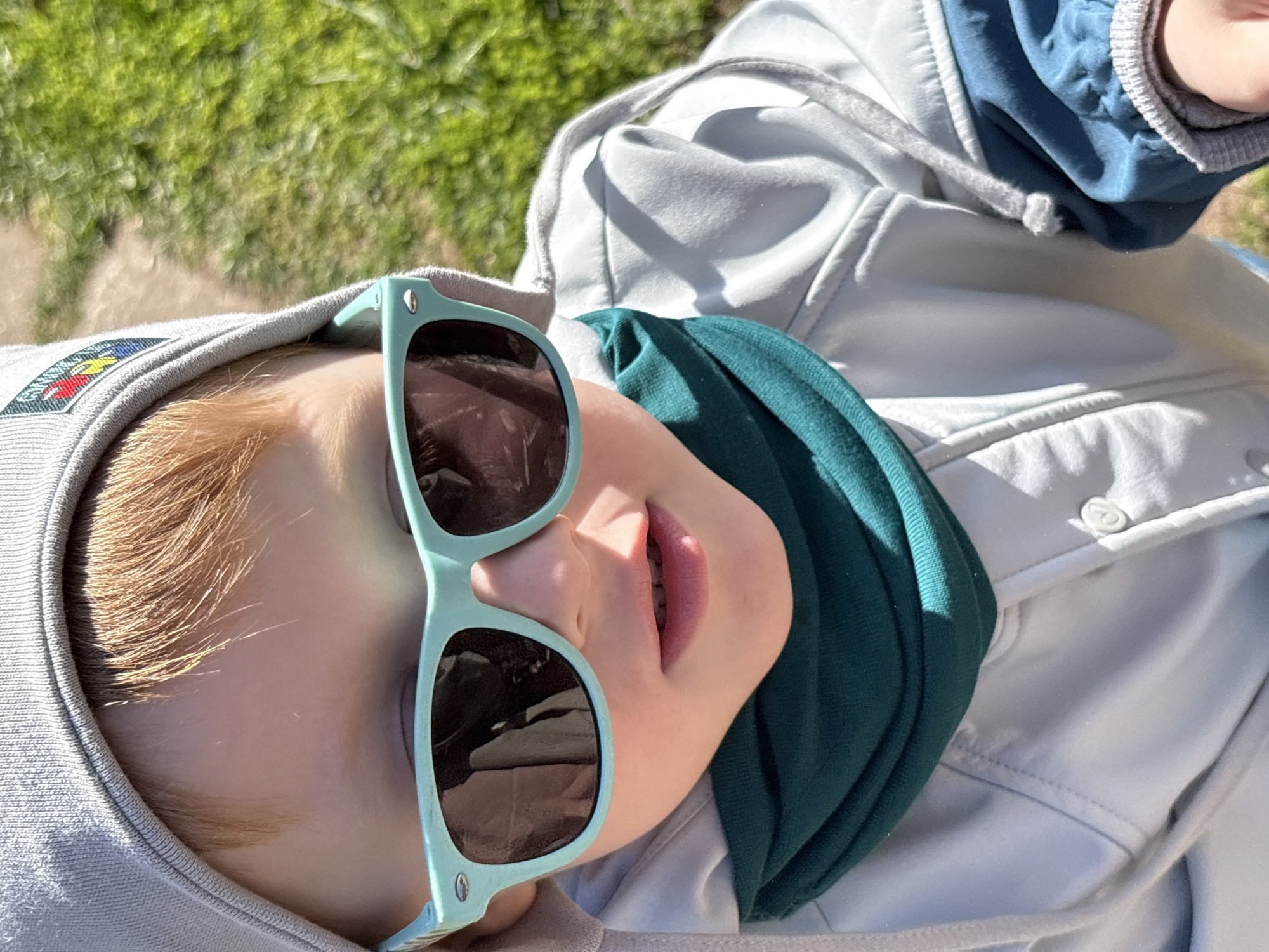 A child wearing light blue sunglasses is the central focus of this eye-level shot, taken outdoors in daylight. The child has fair skin and light-colored hair, and is clad in a grey hooded sweatshirt with a patch on the left side of the hood and a dark teal neck warmer. They are also wearing a white jacket with a button visible on the right side of the frame. The background appears to be a grassy area, suggesting a park or yard setting. The image is slightly rotated, giving the impression that the subject is lying down and looking up at the camera. No specific features or objects are recognizable to place the scene in the city of Italmas, Russia.