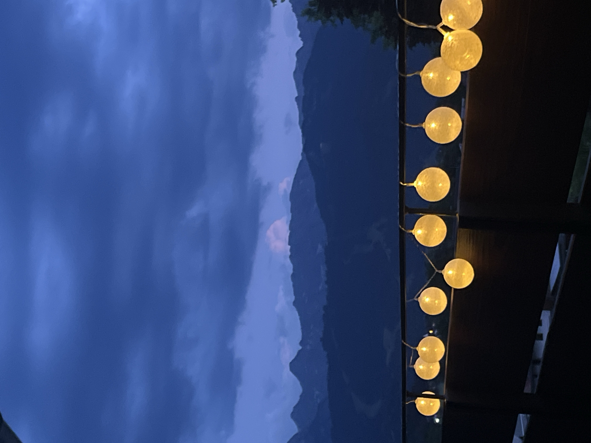 The image depicts a dimly lit outdoor scene, likely at dusk or early evening, overlooking a mountain range. The foreground features a dark, horizontal railing adorned with a string of illuminated, spherical lights. These lights emit a warm, yellow glow. Behind the railing, a dark, wooden structure is visible, possibly a balcony or deck. The middle ground is dominated by the silhouette of a vast mountain range against a deep blue, cloudy sky. The mountains are layered, with darker, closer peaks in shadow and lighter, more distant ones. A faint pinkish hue is present in a cloud formation above one of the peaks. The overall atmosphere is serene and somewhat moody, enhanced by the twilight conditions and the interplay of light and shadow. Given the mountain landscape, the scene could be located in a mountainous region like Garmisch-Partenkirchen, Germany, known for its alpine scenery. There are no people or discernible actions in the image.