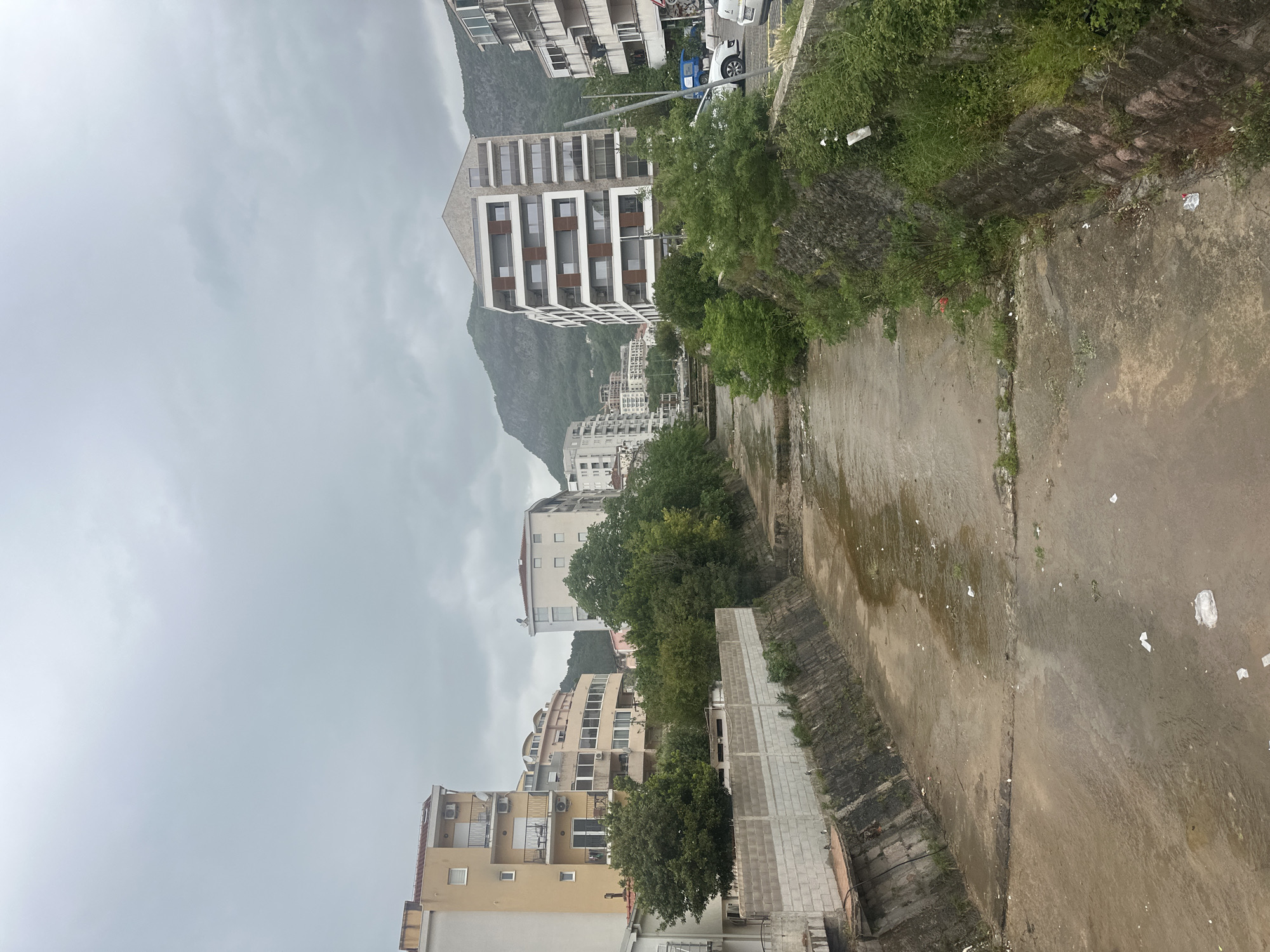 The image depicts an urban scene in Budva, Montenegro, characterized by a concrete drainage canal or riverbed running through the middle of the frame. The canal appears to be mostly dry, with some wet patches and scattered litter. On either side of the canal are embankments, one with vegetation and another with a concrete pathway. Beyond the embankments, residential buildings of various heights and designs are visible. In the background, a mountain rises prominently against an overcast sky. Cars are parked at the top of the image and the buildings are various colours. There are no people visible.