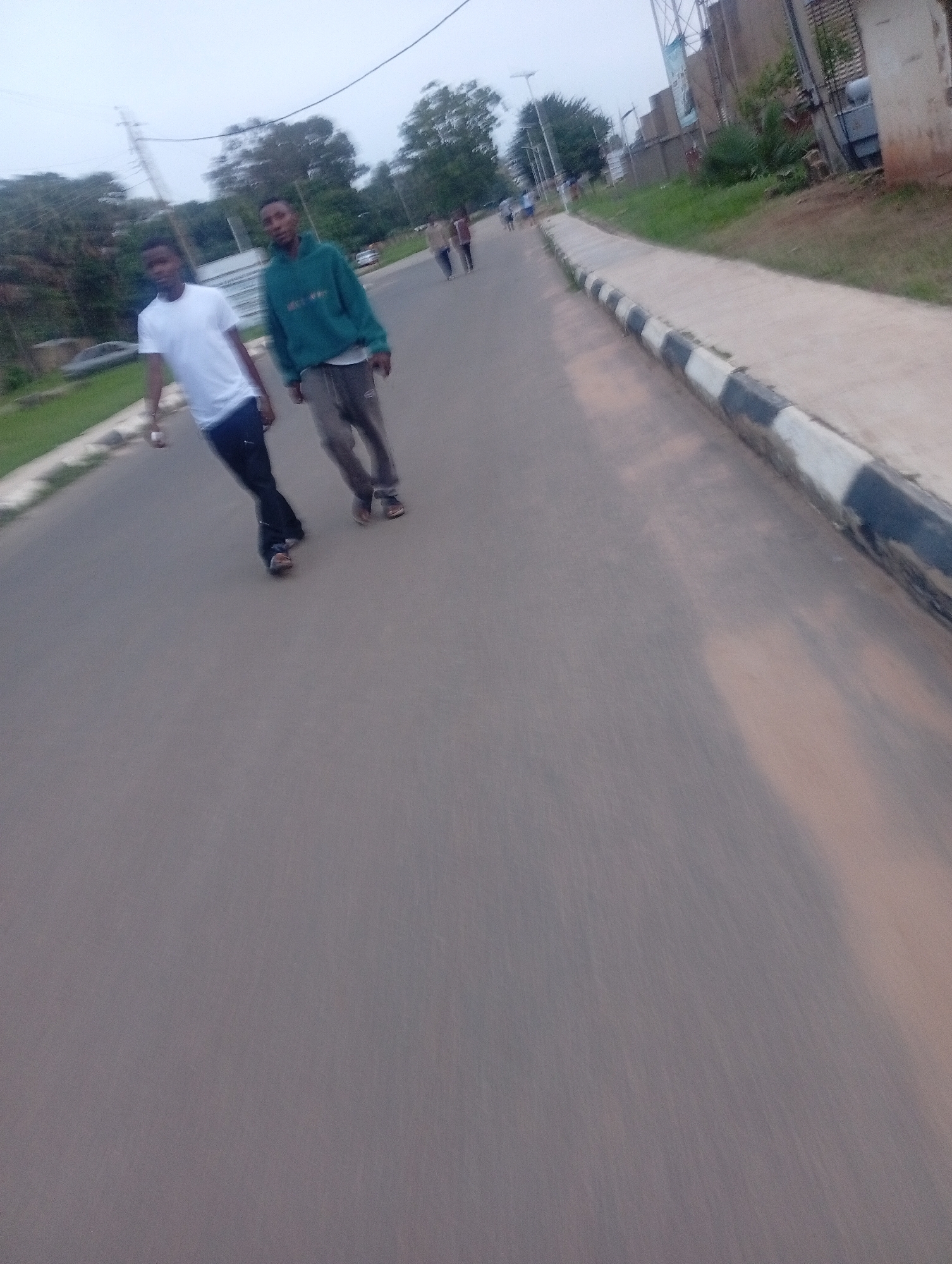 This is a medium-shot, eye-level, wide-angle, outdoor photograph of two young men walking on a paved road. The man on the left is wearing a white t-shirt and dark pants, and is positioned slightly ahead and to the left of the other man. The man on the right is wearing a teal hoodie and grey pants. They are walking towards the camera, and their bodies are angled slightly to the left. The road is asphalt and has a distinct curb with black and white markings on its edge. To the left of the road, there is a grassy area with trees and some structures in the background, including what appears to be a power pole and some buildings. To the right of the road, the pavement continues, and there are more trees and some buildings visible in the distance. The sky is overcast. The overall impression is that of a casual daytime scene, possibly in a residential or semi-urban area. The specific location in Jos, Nigeria, cannot be definitively determined from the visual information alone, but the presence of the distinctive curb and the style of buildings suggest a built-up area within the city.