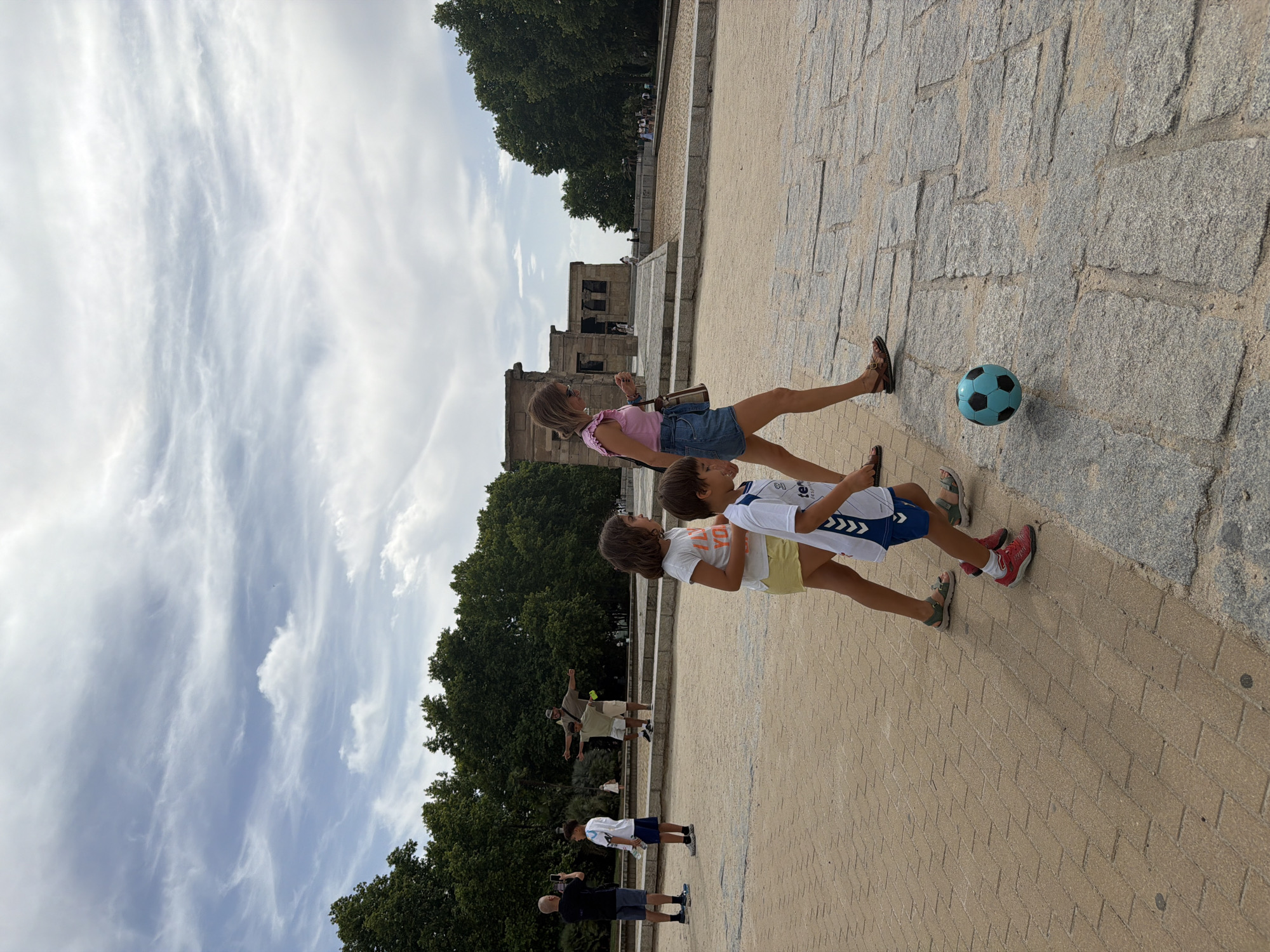 The image depicts the Temple of Debod in Madrid, Spain, under a cloudy sky. In the foreground, a woman in a pink top and denim skirt walks to the right, carrying a brown bag. Beside her, a young boy in a white soccer jersey and blue shorts runs towards a teal soccer ball on the ground. Another child in a yellow skirt and white t-shirt walks to the left, with another child in a white t-shirt and blue shorts behind them. In the background, several people are visible, including a man taking a photo with his phone, and another man posing with his arms outstretched. The temple, an ancient Egyptian structure, is situated on a raised platform with trees and other foliage surrounding it. The ground is paved with stone slabs and cobblestones, with some sandy areas.