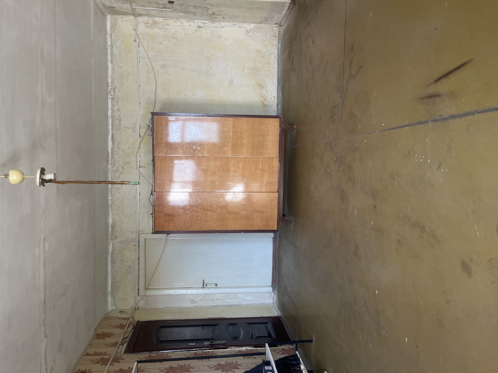 The image depicts an interior scene, likely a room in an apartment in Volzhsky, Russia. The room appears somewhat neglected, with peeling wallpaper and stained walls and ceiling. The flooring is a dark, matte material, possibly linoleum, with visible seams and scuff marks.
Dominating the center of the room is a large, wooden wardrobe with three doors. The wood is a light, polished tone, and the doors have vertical paneling. The wardrobe stands on short, tapered legs.
To the left of the wardrobe is a closed, dark-colored door with ornate paneling and a visible doorknob. Adjacent to this door, and to the left of the frame, is a narrow, black metal shelf unit holding several pairs of shoes.
Further to the left, a portion of patterned wallpaper is visible. A white door with a simple handle is positioned between the dark entrance door and the wardrobe.
The ceiling is textured and stained, with a visible light fixture hanging from a cord. The overall impression is of an older, possibly unoccupied or recently vacated living space. There are no people or animals present in the image. The lighting suggests it is daytime, with natural light reflecting off the wardrobe doors.