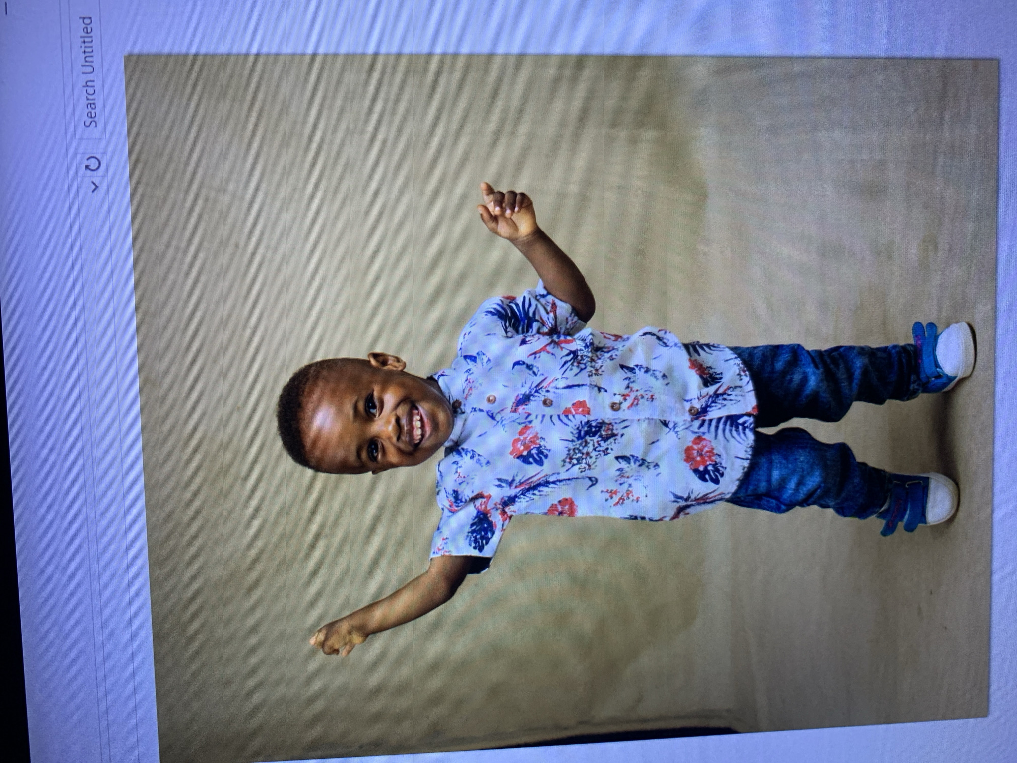 A young Black boy stands facing the camera, his arms outstretched and raised. He is wearing a short-sleeved button-down shirt with a white background and a blue and red floral and feather pattern, dark wash denim jeans, and blue sneakers with white soles and velcro straps. The boy has short, dark hair and a wide smile, revealing his teeth. He is positioned in front of a plain, light brown backdrop. The image appears to be a digital photograph displayed on a computer screen, as indicated by the visible browser interface at the top of the image. No specific location within Gura Top, Nigeria, can be determined from the provided image.