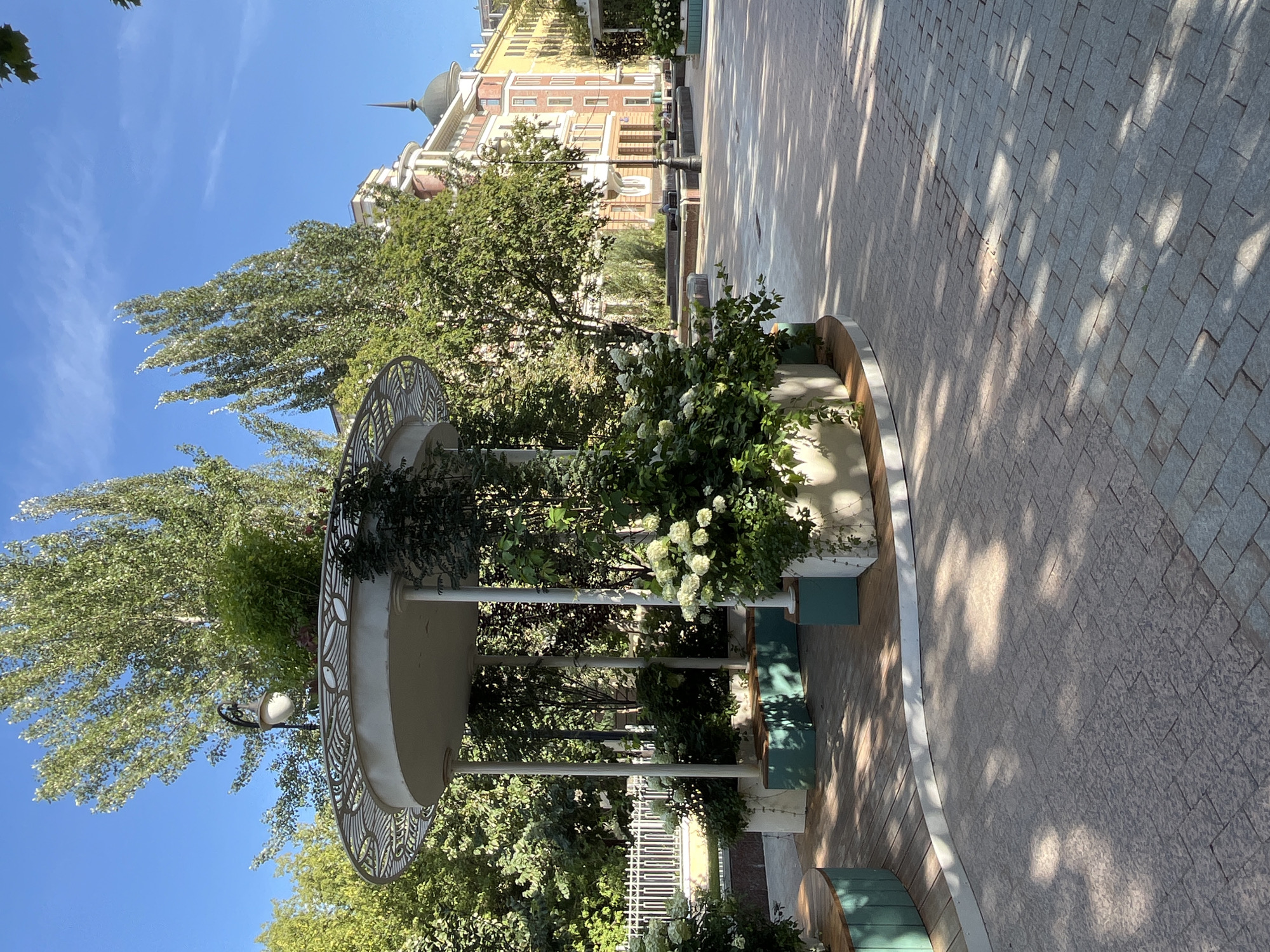 The image depicts a bright, sunny day in a city park or public square in Moscow, Russia. In the foreground, a white, open-air gazebo with decorative metalwork on its roof is surrounded by lush green plants and white hydrangeas. The gazebo has white columns supporting the roof and built-in seating areas with teal-colored panels. The ground in front of the gazebo is paved with interlocking stone tiles, with dappled sunlight creating patterns on the surface.
Behind the gazebo, a wide walkway stretches into the distance, lined with trees and buildings. The buildings are of varying architectural styles, with a prominent multi-story building featuring a red facade, yellow accents, and a dark domed roof with a spire. A black lamppost stands in the middle ground. The overall atmosphere is serene and well-maintained, suggesting a popular public space. There are no people visible in the image.