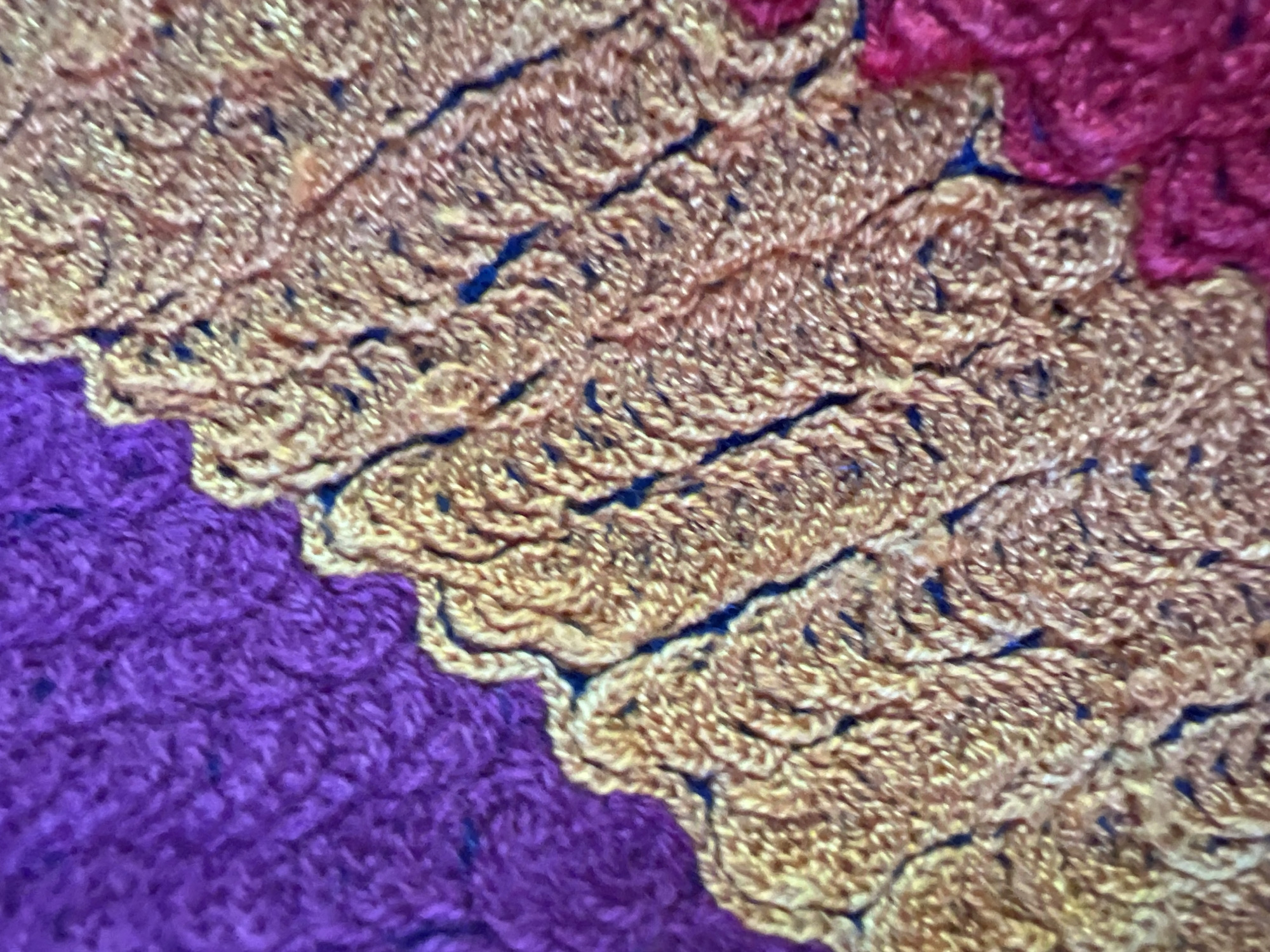The image presents a detailed, close-up view of a knitted or crocheted textile, located in the city of Slivovo, Bulgaria. The fabric displays three distinct color sections: a rich purple in the upper left quadrant, a prominent golden-yellow and orange blend occupying the central and upper-right portions, and a vivid reddish-pink hue in the lower right. The textile exhibits a pronounced texture, characterized by visible loops and interwoven strands of yarn, forming a raised, relief-like surface. Irregular, wavy lines, appearing as dark, shadowed seams or deeper stitches, delineate the transitions between these color blocks, suggesting a structured pattern within the fabric. No individuals, further objects, or specific environmental contexts beyond the textile itself are discernible within the frame. The focus is entirely on the intricate pattern and tactile qualities of the multi-colored yarn.
