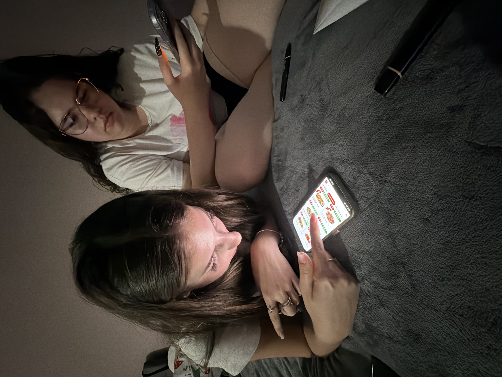 The scene depicts two young adult females indoors, likely within a residential setting in the city of Burgas, Bulgaria. Both individuals are positioned on a dark grey, textured blanket or comforter. The ambient lighting is dim, with the primary light source appearing to be the illuminated screens of their respective smartphones.
The female in the foreground, located on the left, is lying prone with her upper body slightly elevated. She has long, dark brown hair and is wearing a light-colored, short-sleeved t-shirt. Her attention is focused downward on her smartphone, the screen of which displays an application interface featuring various food items, potentially pizzas, along with numerical values and status indicators (e.g., green checkmarks, red 'x' marks). Her right index finger is in contact with the screen, while her left hand, adorned with multiple rings, gestures towards the display.
The second female, situated in the background on the right, is seated or leaning back. She has dark hair and wears gold-rimmed glasses. Her attire consists of a white short-sleeved t-shirt with a subtle pink graphic print and dark-colored shorts or undergarments. She holds a silver-toned smartphone in her right hand, her fingernails displaying a distinct dark base with orange and yellow tips. Her gaze is directed towards the left, appearing to observe the area in front of her.
Between the two individuals, a black cylindrical object with a gold band lies on the grey blanket. A white piece of paper or fabric is also visible further to the right on the blanket. The immediate background shows a plain, light-colored wall.