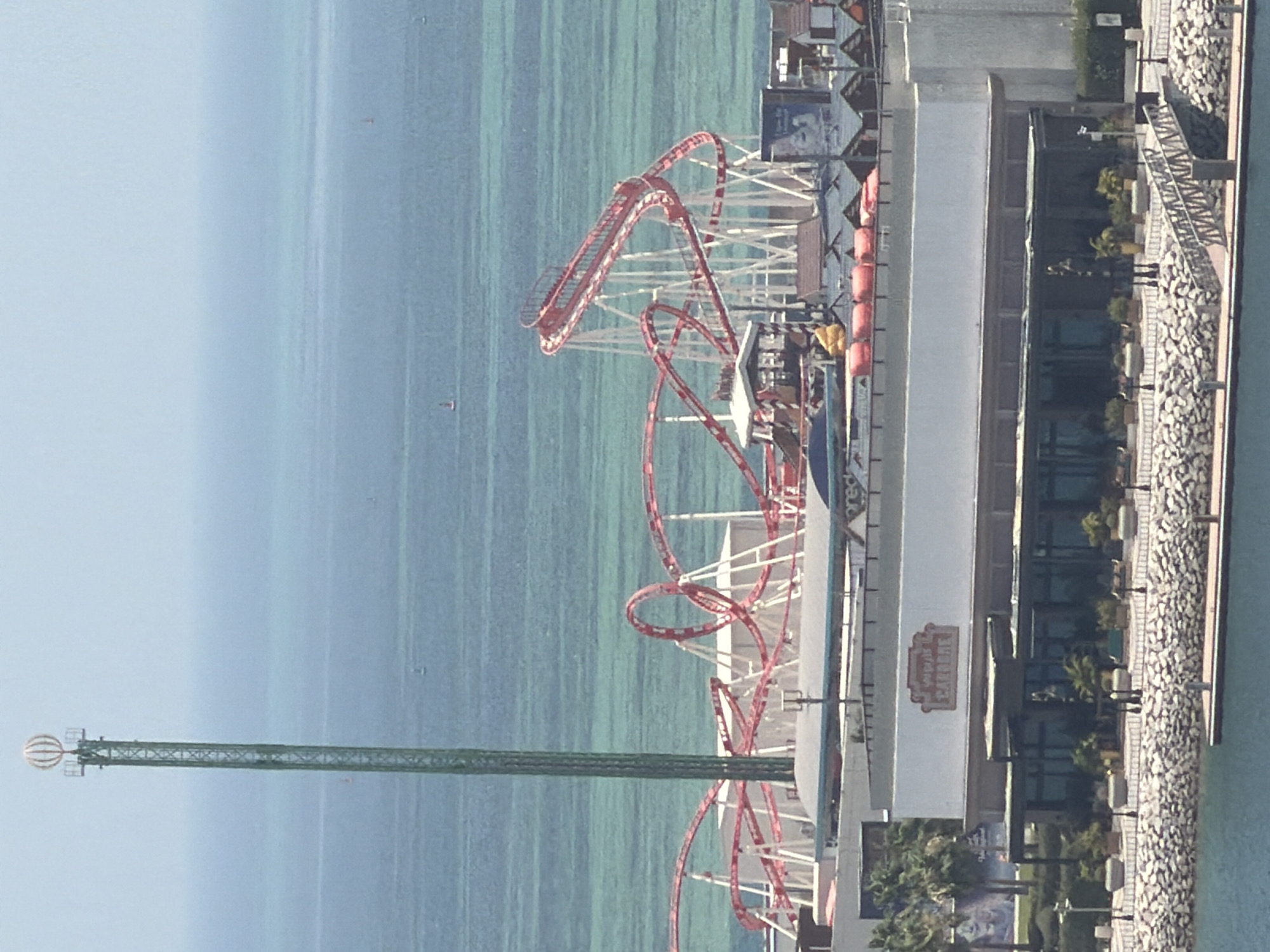 The image shows an amusement park with a prominent red and white roller coaster in the foreground. To the left of the roller coaster is a tall, slender green tower topped with a spherical, segmented structure. The amusement park appears to be situated near the ocean, with a large expanse of blue-green water extending to the horizon. In the distance, several small boats are visible on the water. Below the roller coaster, there is a building with a sign that reads "CARIBE". The building is modern in design, with large glass windows. The area in front of the building is paved with stone and lined with potted plants and lampposts. The scene suggests a coastal amusement park or resort.