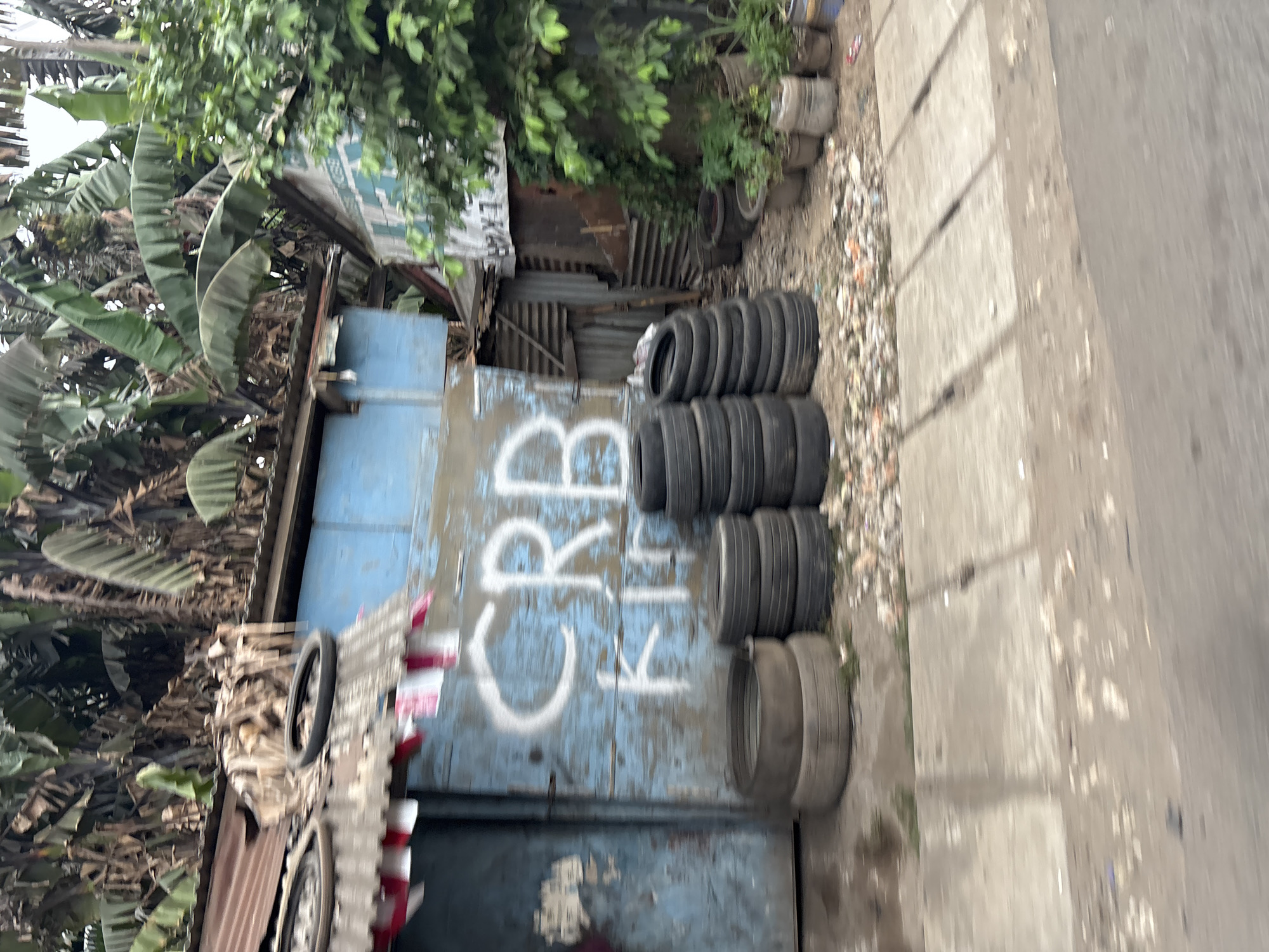 The image depicts a scene in Bekasi, Indonesia, featuring a small, weathered blue building with corrugated metal roofing. The building's facade is adorned with graffiti that appears to read "CRB KIB." In front of the building, a pile of old tires is stacked haphazardly, with two tires also placed on top of the roof. Lush green foliage, likely banana trees, surrounds the structure. The ground in front of the building is paved with concrete slabs and shows signs of debris. The overall impression is one of a modest, potentially informal commercial establishment or residence in an urban environment.