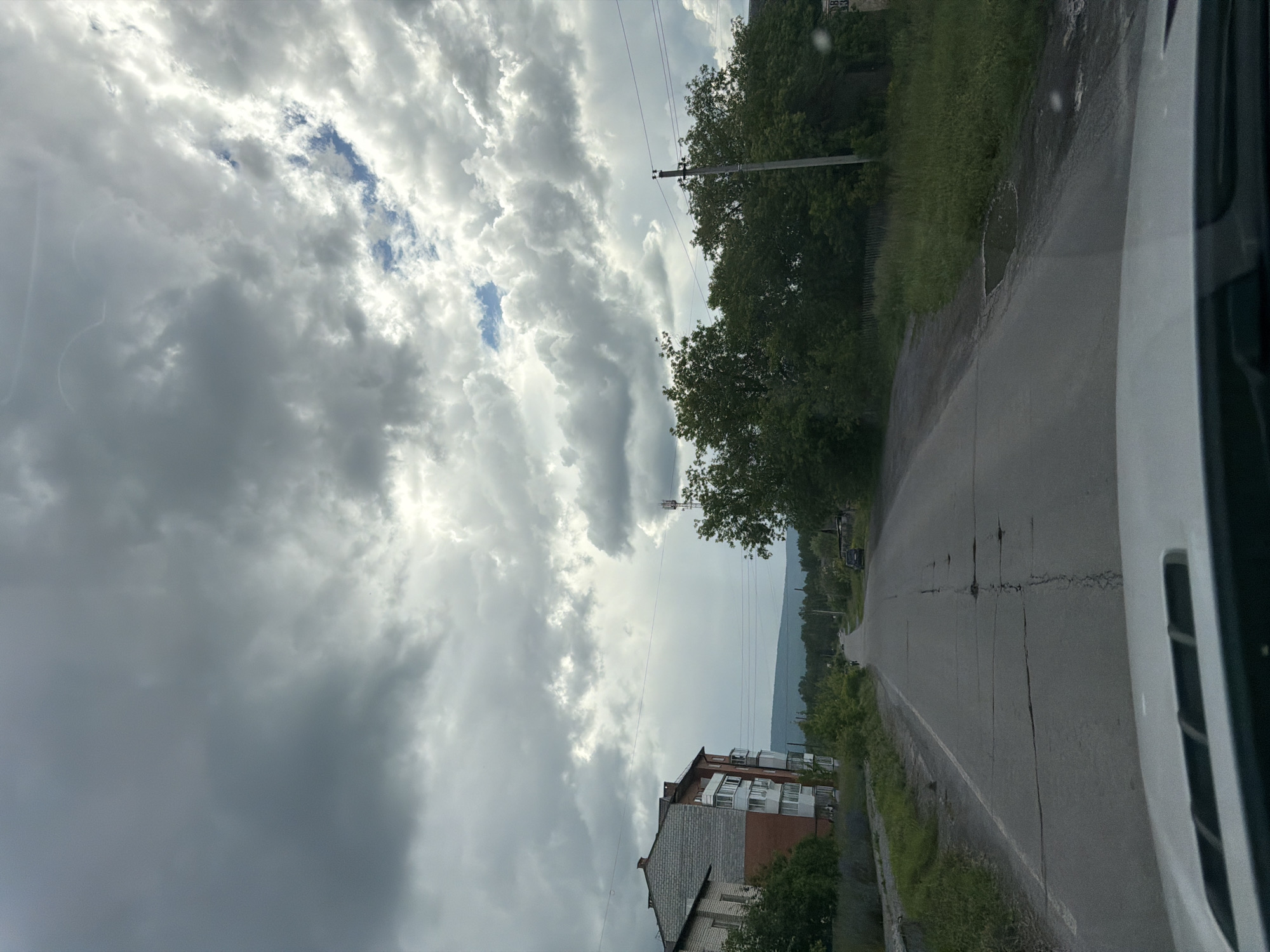 The image captures a view from a vehicle driving on a cracked asphalt road in Mezhevoy, Russia. The sky is overcast with dramatic, heavy gray clouds interspersed with patches of bright sunlight, suggesting variable weather. To the left of the road, a multi-story brick and concrete apartment building stands, with balconies visible on its facade. On the right side of the road, dense green trees line the foreground and extend into the middle ground, with a telephone pole and antenna also present. In the distance, a range of dark green hills forms the horizon, with a few scattered houses and more trees visible along the roadside. The road itself shows signs of wear, with visible cracks and potholes. The scene depicts a rural or semi-urban environment on what appears to be a somewhat gloomy day.