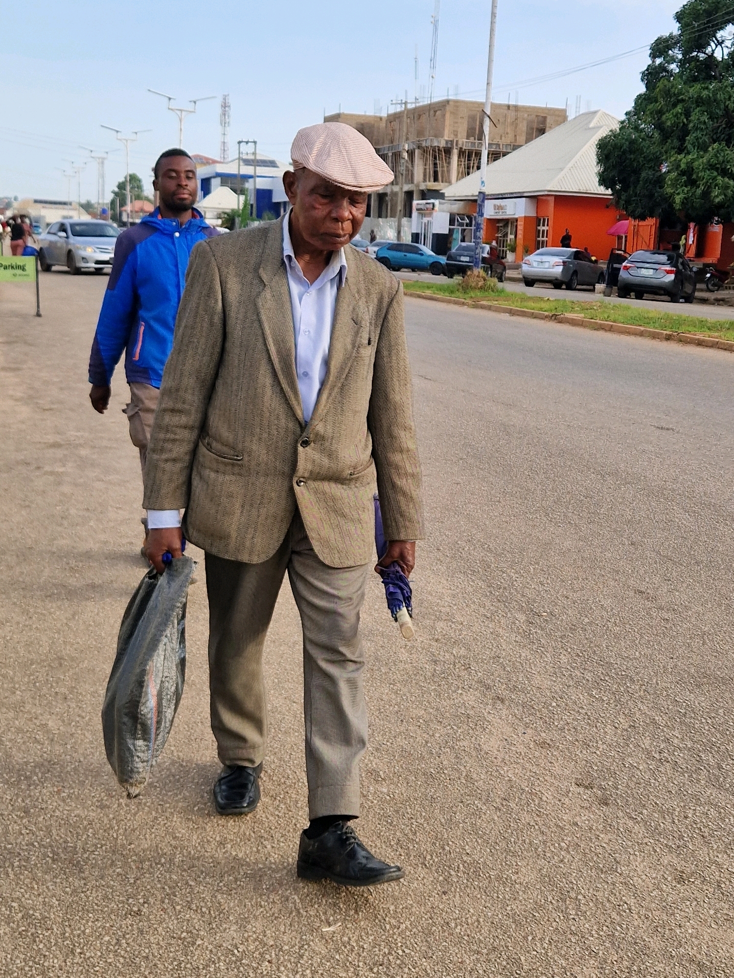 The image captures a street scene in Jos, Nigeria, under a clear sky. In the foreground, an elderly Black man wearing a checkered cap, a light blue collared shirt, a textured brown jacket, and grey trousers walks on a paved road, carrying a black plastic bag in his left hand and a closed blue umbrella in his right. To his left and slightly behind him, a younger Black man in a blue jacket and grey pants walks in the same direction. The background features a street with parked cars, buildings under construction, a small orange building with a white roof, and a large green tree. The road surface is asphalt, and there is a narrow strip of grass along the side of the road. The lighting suggests it is daytime, likely late afternoon.