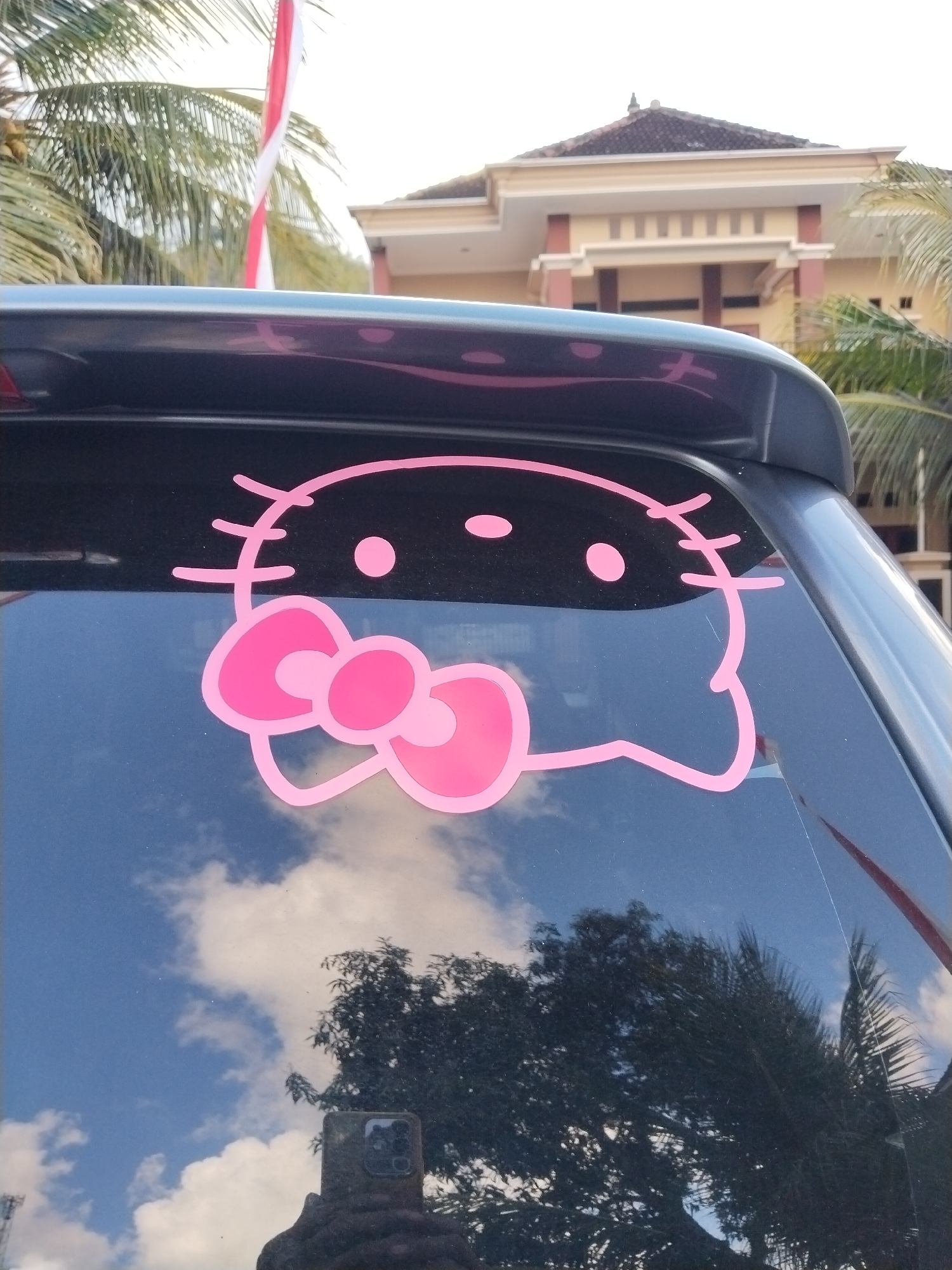 The image shows a close-up view of the rear window of a dark-colored car, featuring a large pink Hello Kitty sticker. The sticker is centrally placed on the window and depicts Hello Kitty's face with her signature bow. Reflections on the car window reveal a bright blue sky with white clouds, trees, and the facade of a building. In the background, a red and white striped flag is visible, along with palm trees and the roof of a building with terracotta tiles. The reflection also captures a person holding a smartphone, likely taking the photograph. The scene appears to be outdoors, possibly in a residential or public area. Based on the context provided, this image was taken in the city of Karanggandu, Indonesia.