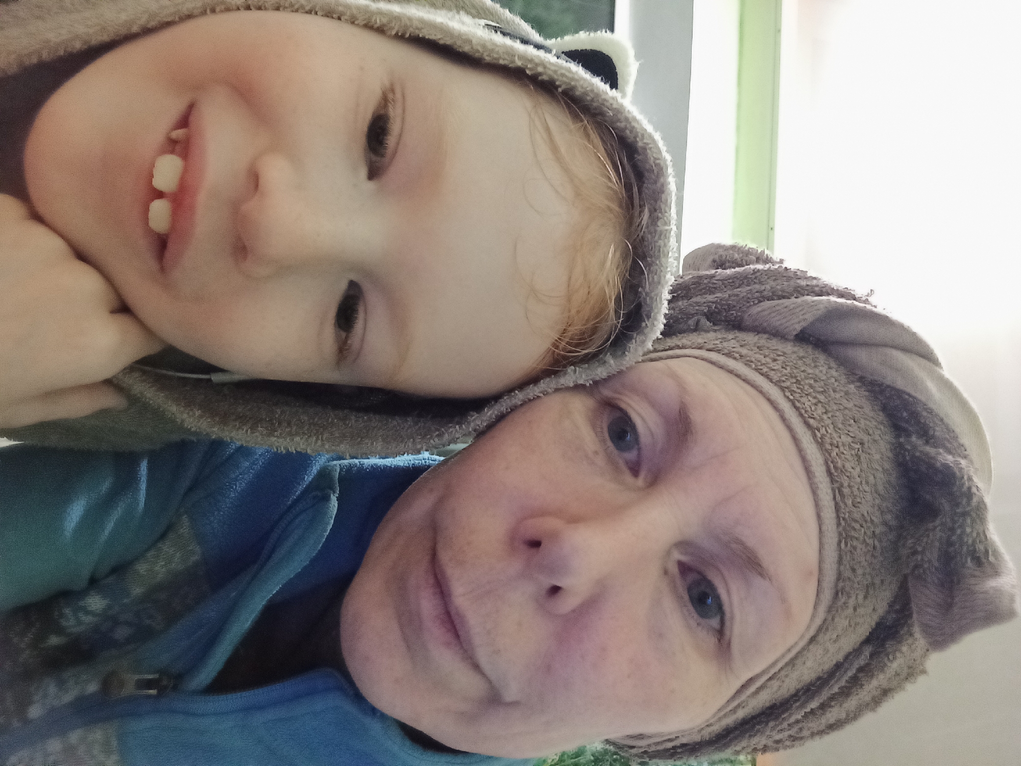 A selfie features a child with red hair and a woman. The child is wearing a grey hooded robe with bear ears and is looking at the camera with a slight smile, propping their chin on their hand. The woman, partially visible to the right, is wearing a grey towel wrapped around her head and a blue jacket. Both subjects have their faces close to the camera, suggesting a personal moment. The background is blurred but appears to be an indoor setting with some light from a window. The image appears to have been taken indoors in Blagoveshchensk, Russia.