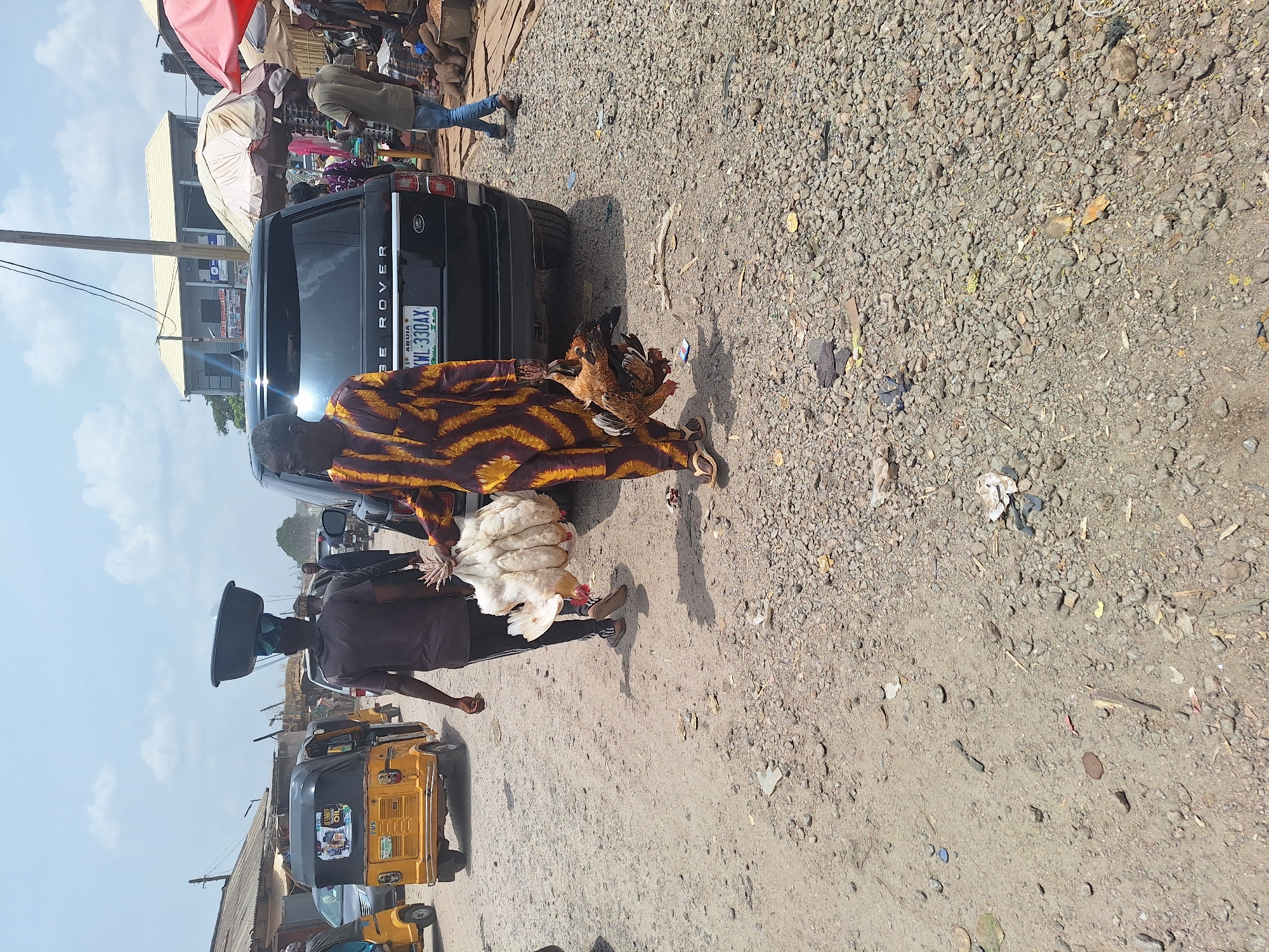 The image shows a busy scene on what appears to be a road or street in Maiburiji, Nigeria. In the center of the frame, a person wearing a striped yellow and brown garment carries brown chickens in one hand. Next to this person, another individual is carrying a load of white chickens. Behind the people and chickens, there is a black Range Rover SUV with a Nigerian license plate. A man wearing a dark shirt is carrying a black container on their head. A yellow "Keke Napep" (tricycle taxi) is in the lower left. The ground is gravelly and dusty, indicative of an unpaved or poorly maintained road surface. There are various pedestrians, stalls, and buildings visible in the background, suggesting a market or commercial area.