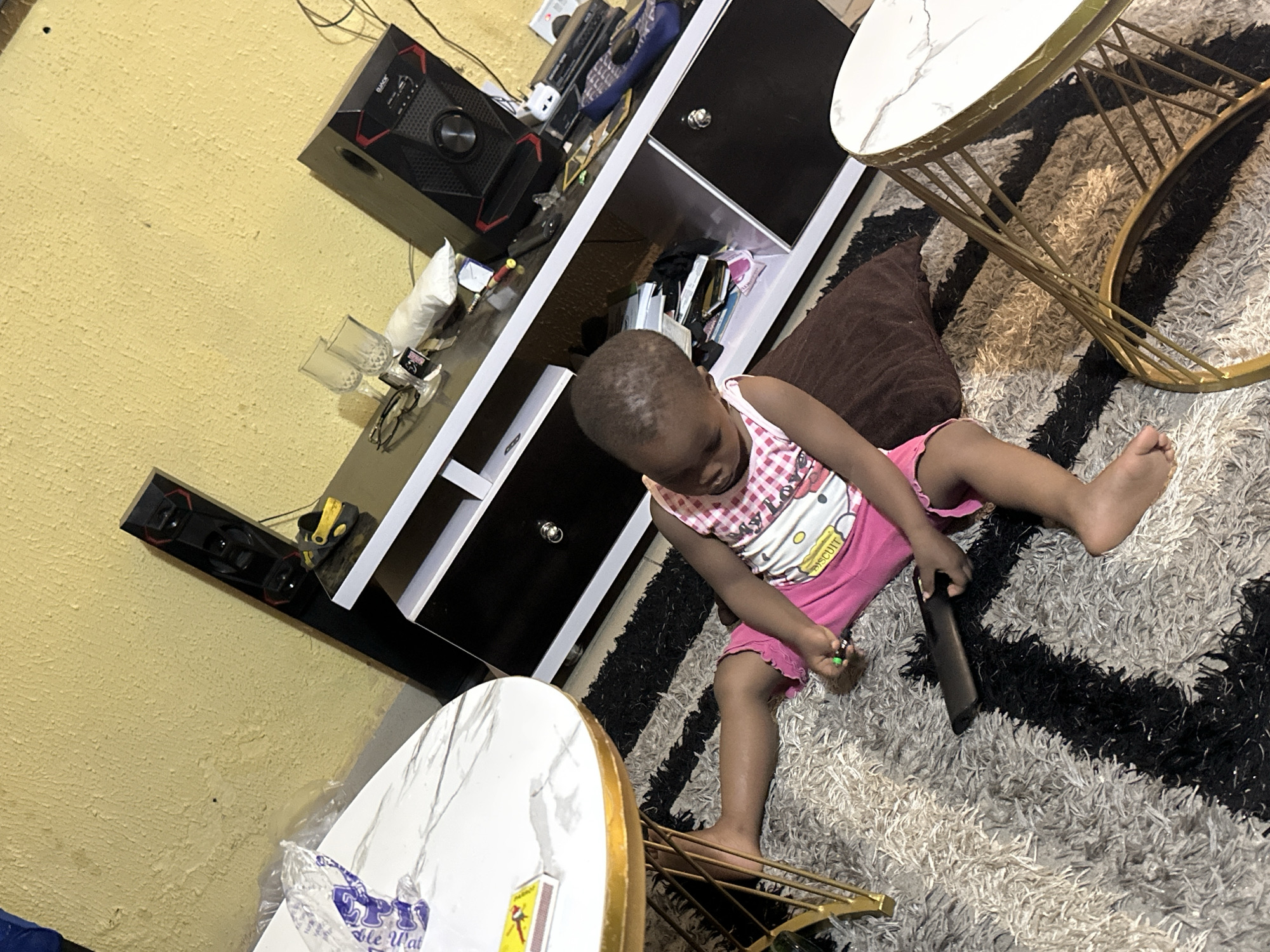 A toddler, wearing a pink and white checkered top with a Hello Kitty graphic and pink shorts, sits on a black and white striped rug in what appears to be a living room in Narayi, Nigeria. The child is holding a dark-colored object, possibly a remote control, and a green pen in their hands. The child's legs are spread apart, and their feet are bare. In the background, a dark entertainment unit holds various items, including speakers, glasses, and electronic devices. A yellow wall with a textured surface is visible behind the entertainment unit. To the left of the scene, a white and gold marble-top coffee table is partially visible. To the right, another similar coffee table with a marble top and gold legs is also present. The lighting suggests an indoor environment.