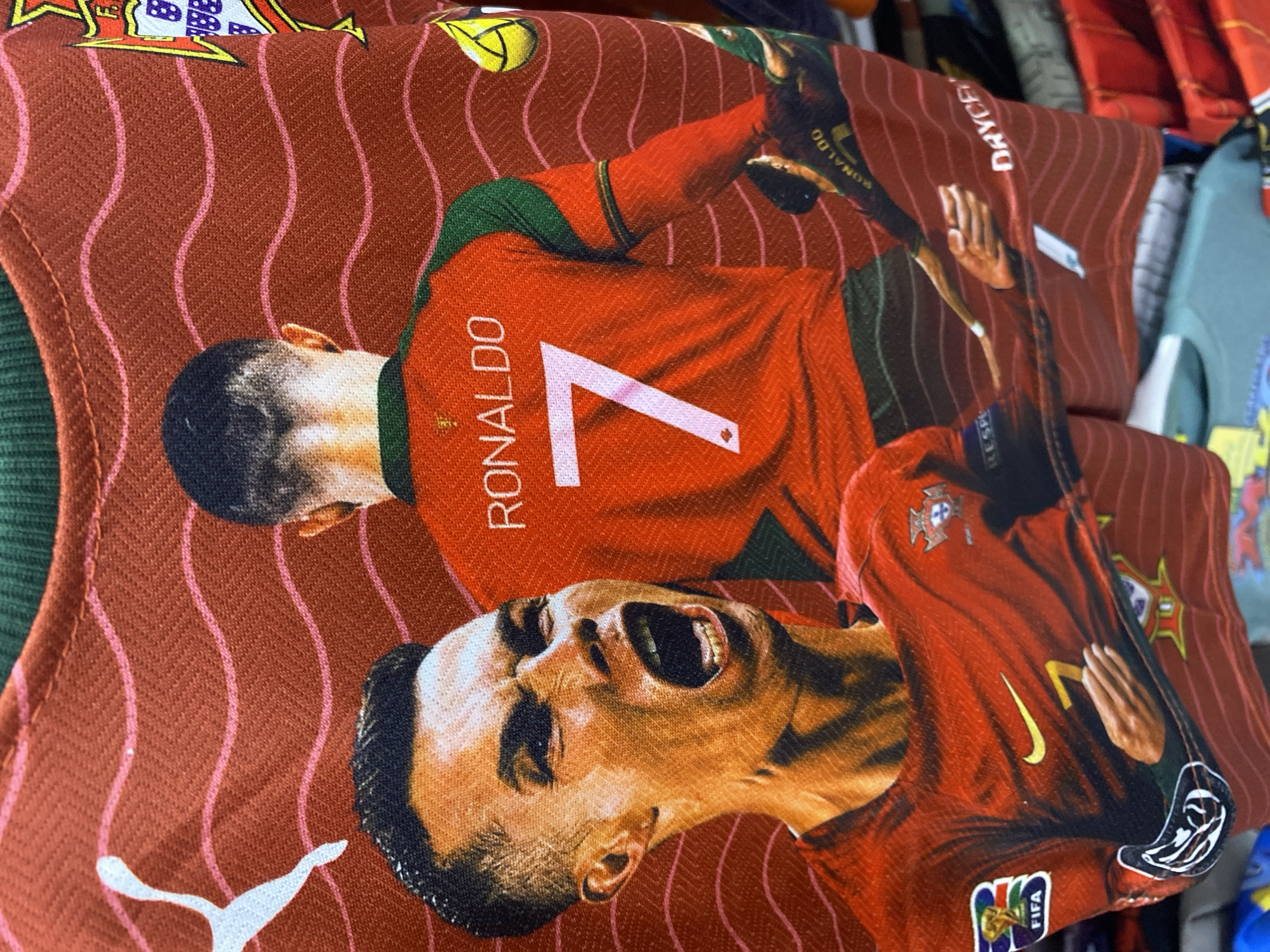 The image shows a close-up of a red t-shirt featuring a graphic of Cristiano Ronaldo. Ronaldo is depicted on the front of the shirt with his mouth open in a shout, and on the back, showing his jersey number "7" and the name "RONALDO". The t-shirt has wavy red and pink lines in the background and features logos such as Puma, Nike, FIFA, and the Portuguese national team crest. The shirt appears to be on display in a retail setting, with other t-shirts visible in the background. The scene is a retail store in Banda Aceh, Indonesia.