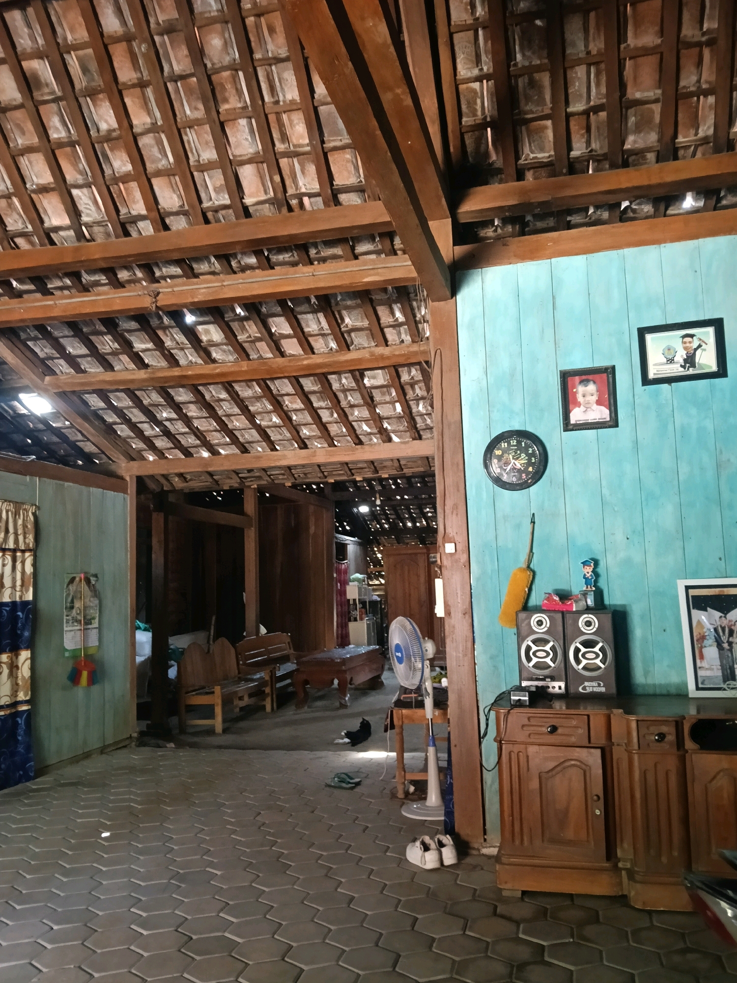 The image depicts the interior of a room with a wooden beam and tile roof structure. The walls are painted turquoise. On the wall, there are two framed photographs, a circular clock, and a yellow duster. Below the clock, there are two speakers and a wooden cabinet with drawers and doors. In the foreground, a white oscillating fan is positioned on a stand. A pair of white shoes is placed on the tiled floor. To the left, there is a curtain with a patterned design. In the mid-ground, there is wooden seating and further into the room, more wooden structures and furniture are visible. A black cat is lying on the floor in the mid-ground. The floor is tiled with hexagonal grey tiles. The scene appears to be located in Nglungger, Indonesia, given the architectural style and common household items.