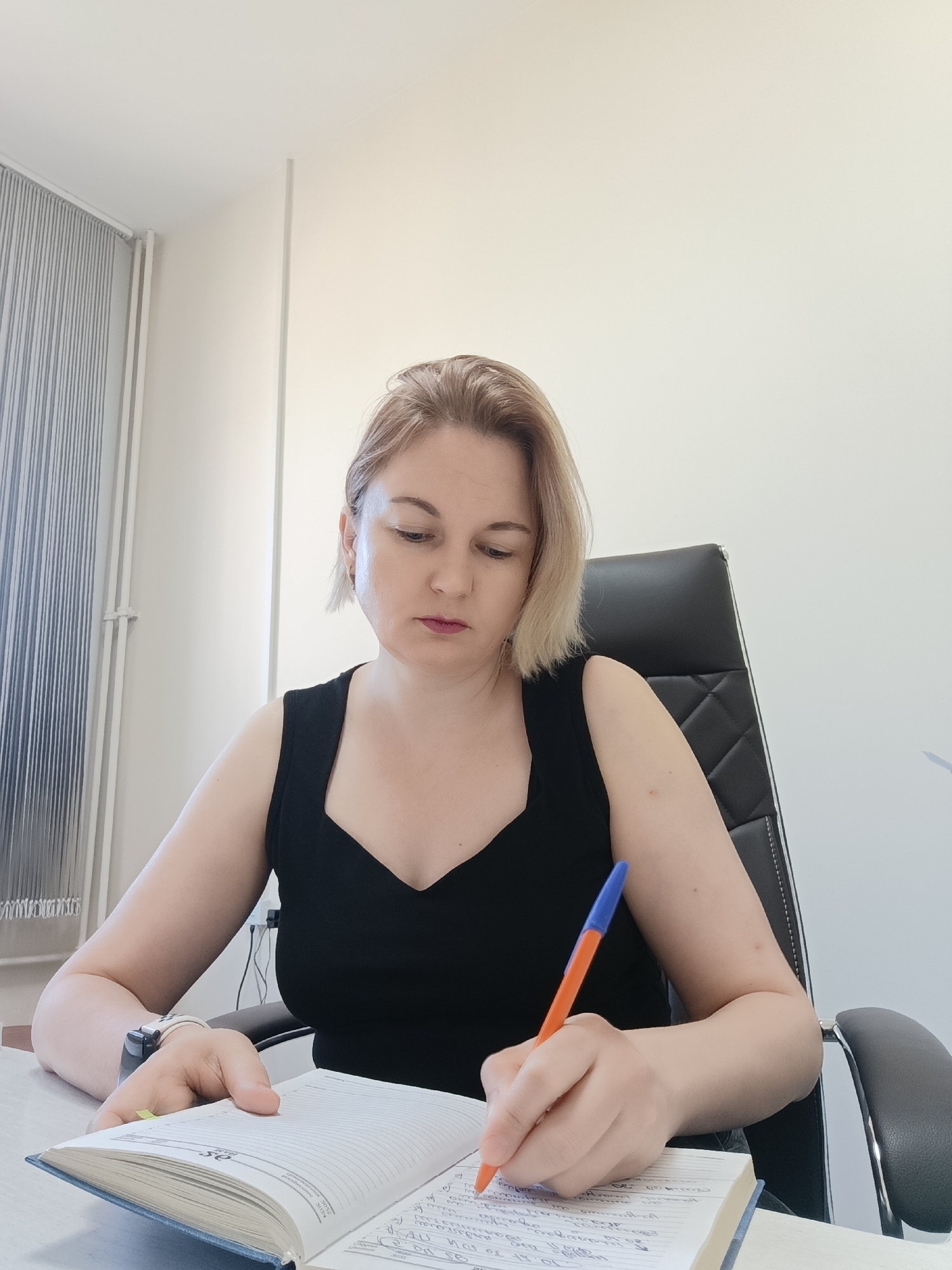A blonde woman sits at a desk, writing in a notebook with a pen. She wears a sleeveless black top and a watch on her left wrist. The notebook is open on the desk in front of her, filled with handwritten notes. She is sitting in a black office chair with a quilted back. The background is a plain white wall with a window covered by blinds to the left. The location is Марковский, Russia.