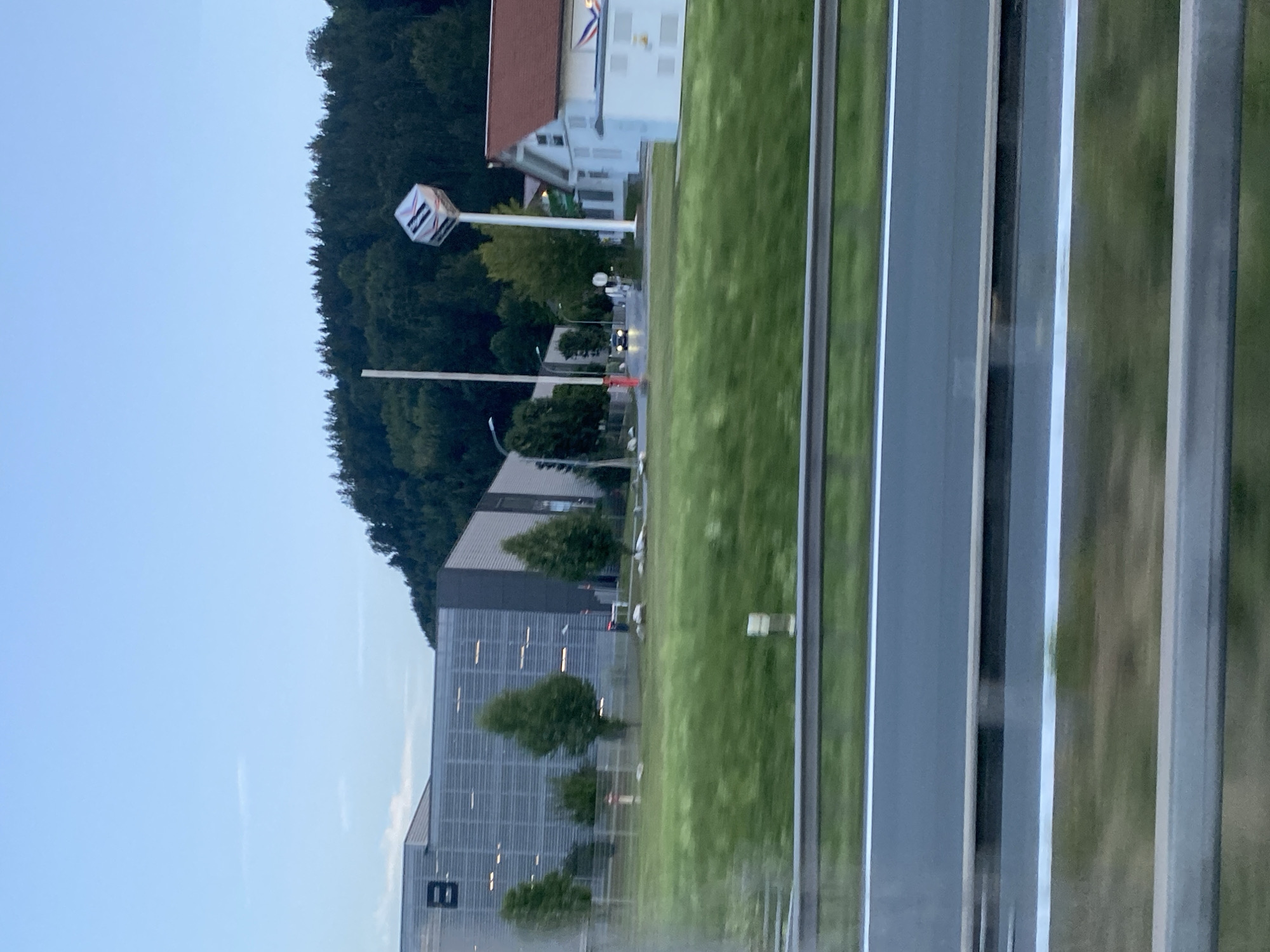 The image depicts a roadside view, likely from a vehicle traveling on a highway, showing a business complex and a forested hillside in the background. On the left is a large, modern building with a prominent "B" logo. Adjacent to it, a grassy area with scattered trees is visible. To the right, a white building with a red-tiled roof is present, along with a tall signpost displaying a logo, and what appears to be a gas station or service area. A car with its headlights on is driving on a road in the mid-ground. The sky is a pale blue, indicating it's either early morning or late evening. The foreground shows a highway with white lane markings, separated by guardrails from the scene behind. The specific location of Siegsdorf, Germany, is not directly evident from visual cues within the image itself.