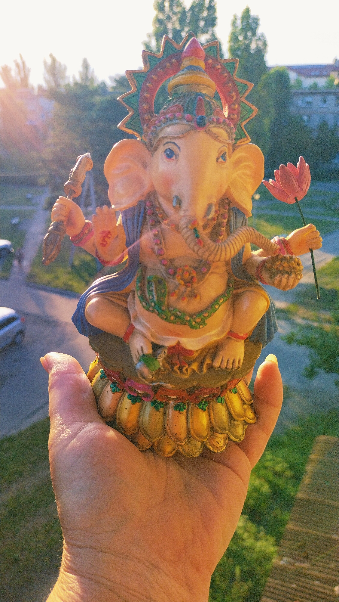 A hand holds a colorful statue of Ganesh, the Hindu deity with an elephant head, against an outdoor urban background. The statue, detailed with multiple arms and adorned with necklaces and a headdress, sits on a lotus-shaped base. Ganesh holds a pink lotus flower in its upper right hand and a small item, possibly a sweet, in its lower right hand. In the background, trees and buildings are visible, suggesting a city environment. The scene is illuminated by sunlight, creating lens flare and casting shadows. The image was captured in Horishni Plavni, Ukraine.
