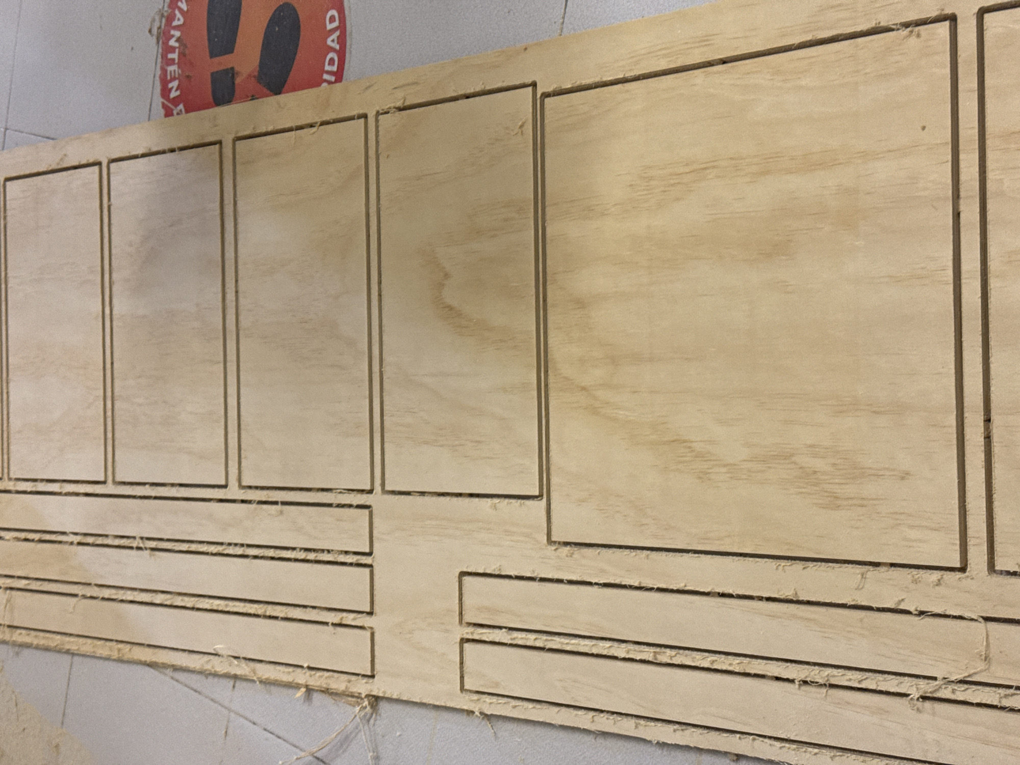 The image shows a sheet of light-colored plywood that has been cut with a CNC router or similar tool. The cuts form a pattern of recessed rectangles and horizontal slots. The edges of the cuts are somewhat frayed, suggesting the wood may be of lower quality or the cutting tool was not perfectly sharp. The wood is resting on a light gray or white surface, likely a workbench or countertop. In the upper left corner of the image, a portion of a round sign with the words "MANTEN" and "IDAD" is visible. Based on the location provided, this image appears to have been taken indoors in San Bernardino Tlaxcalancingo, Mexico. There are no people visible.