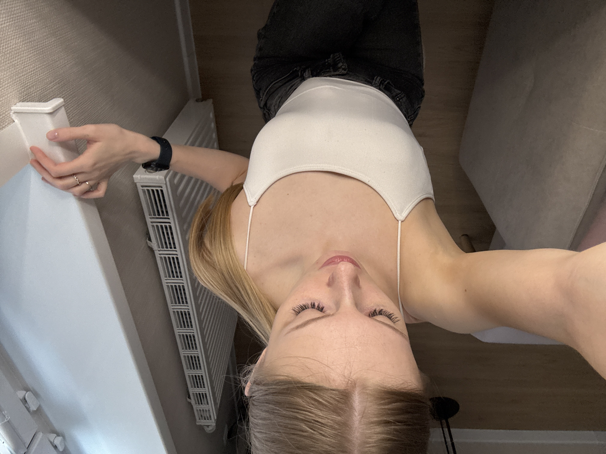 A low-angle, selfie-style photograph shows a blonde woman in a white tank top and dark denim jeans. She is standing indoors, her head pointing towards the bottom of the frame, giving a view up her body. Her eyes are open, and her hair is parted in the middle. She is wearing a black smartwatch on her left wrist and rings on the fingers of her left hand. A white radiator is visible to her left, and another white surface is partially visible behind her on the left. A beige wall is visible in the top left corner. A beige sofa is partially visible on the top right. Based on visual analysis of the scene, there are no explicit indicators of the location being in Perm, Russia.