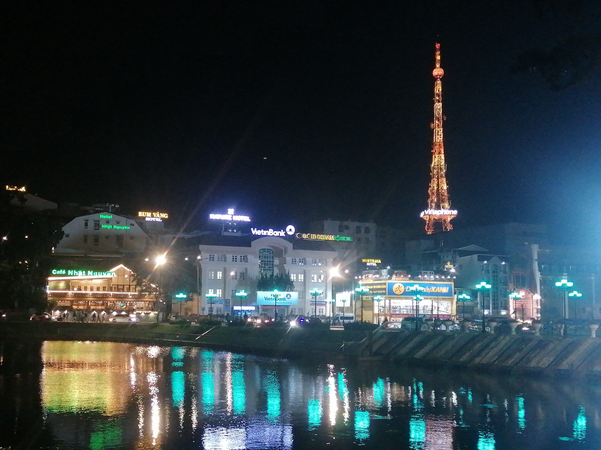 This is a night view of Da Lat, Vietnam, featuring a riverfront scene with several prominent buildings and a tall telecommunications tower in the background. The river reflects the lights of the buildings and street lamps.
Buildings along the riverfront display various signs, including "Café Nhật Nguyên," "VietinBank," "GOLD DREAM," "WISTERIA HOTEL," and "Đáy XANH." A tall, illuminated telecommunications tower, branded "Vinaphone," stands out against the dark sky. Streetlights line the riverbank, and there are some people visible along the promenade.