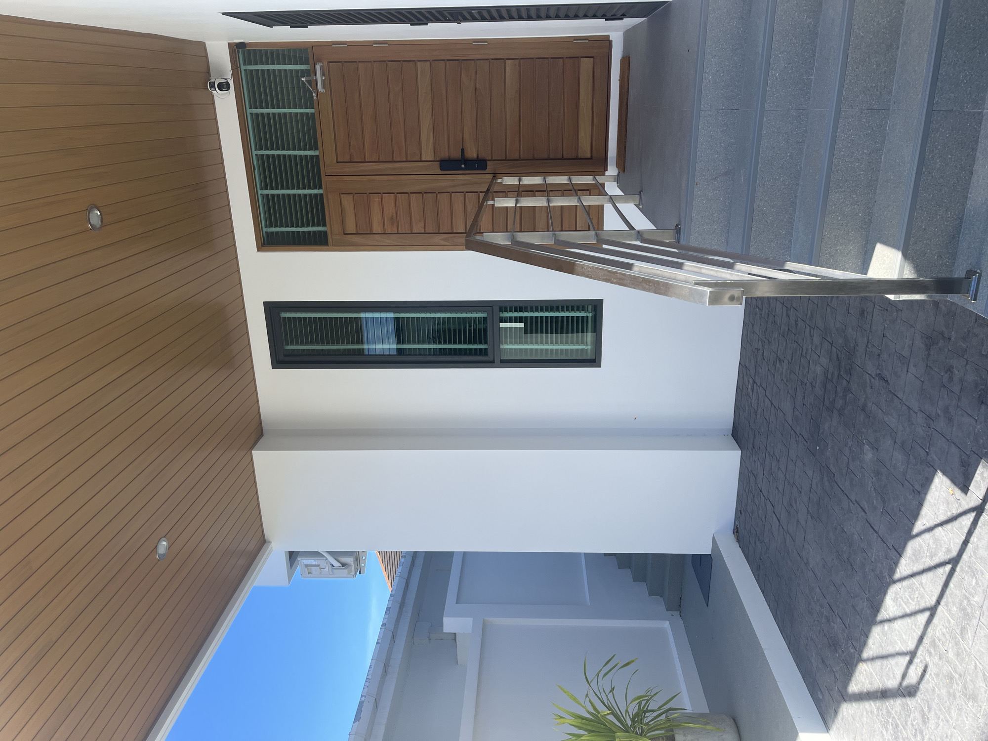 The image depicts the exterior of a modern house located in Rawai, Thailand. The scene is dominated by a clean, white facade with a wooden-paneled ceiling covering an entryway. A set of gray tiled stairs with a stainless steel handrail leads up to a pair of substantial wooden double doors. To the left of the doors, a tall, narrow window with dark framing allows light to filter through louvred blinds. A security camera is visible mounted on the white wall above the doors. The ground in front of the stairs is paved with dark, patterned concrete tiles, where the shadows of an unseen structure are cast. To the left, a walkway made of the same dark tiles leads towards a lower level where a green potted plant stands near a set of outdoor stairs. The overall impression is one of a well-maintained, contemporary residence under a clear blue sky.