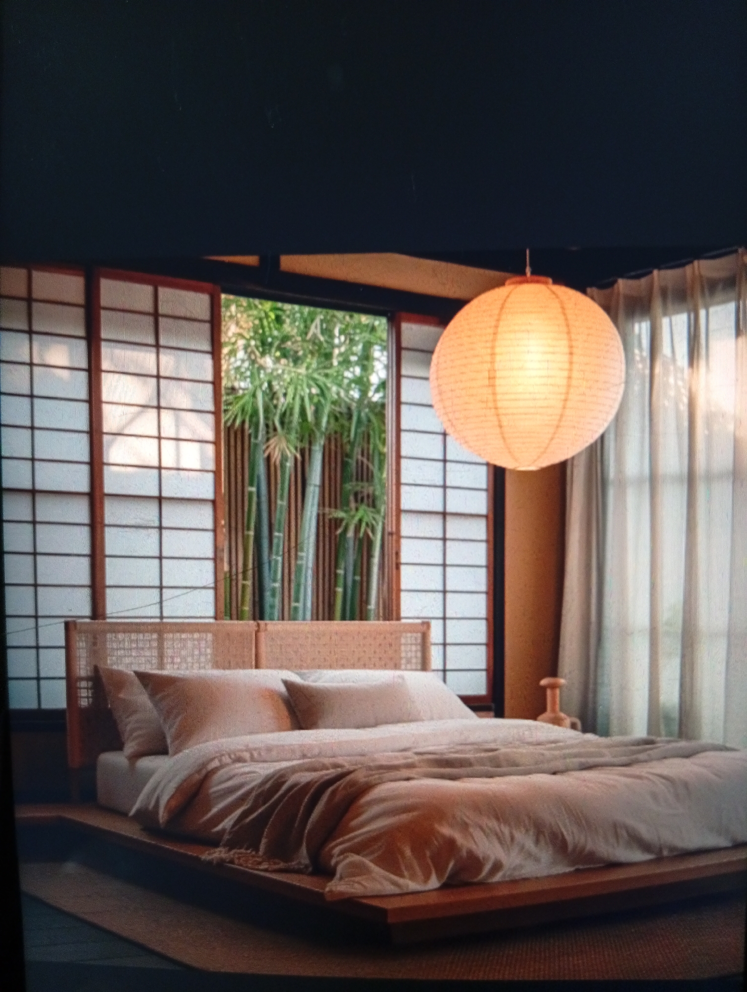 The image depicts a bedroom interior with an Asian-inspired aesthetic. The room features a low platform bed with a light-colored comforter and pillows. Behind the bed, there is a headboard with a woven design. A large paper lantern hangs from the ceiling, providing ambient light. The room has shoji-style windows with a view of bamboo plants outside. A sheer curtain hangs to the right of the windows. The location in Dutsen Alhaji, Nigeria, is not identifiable from the image.