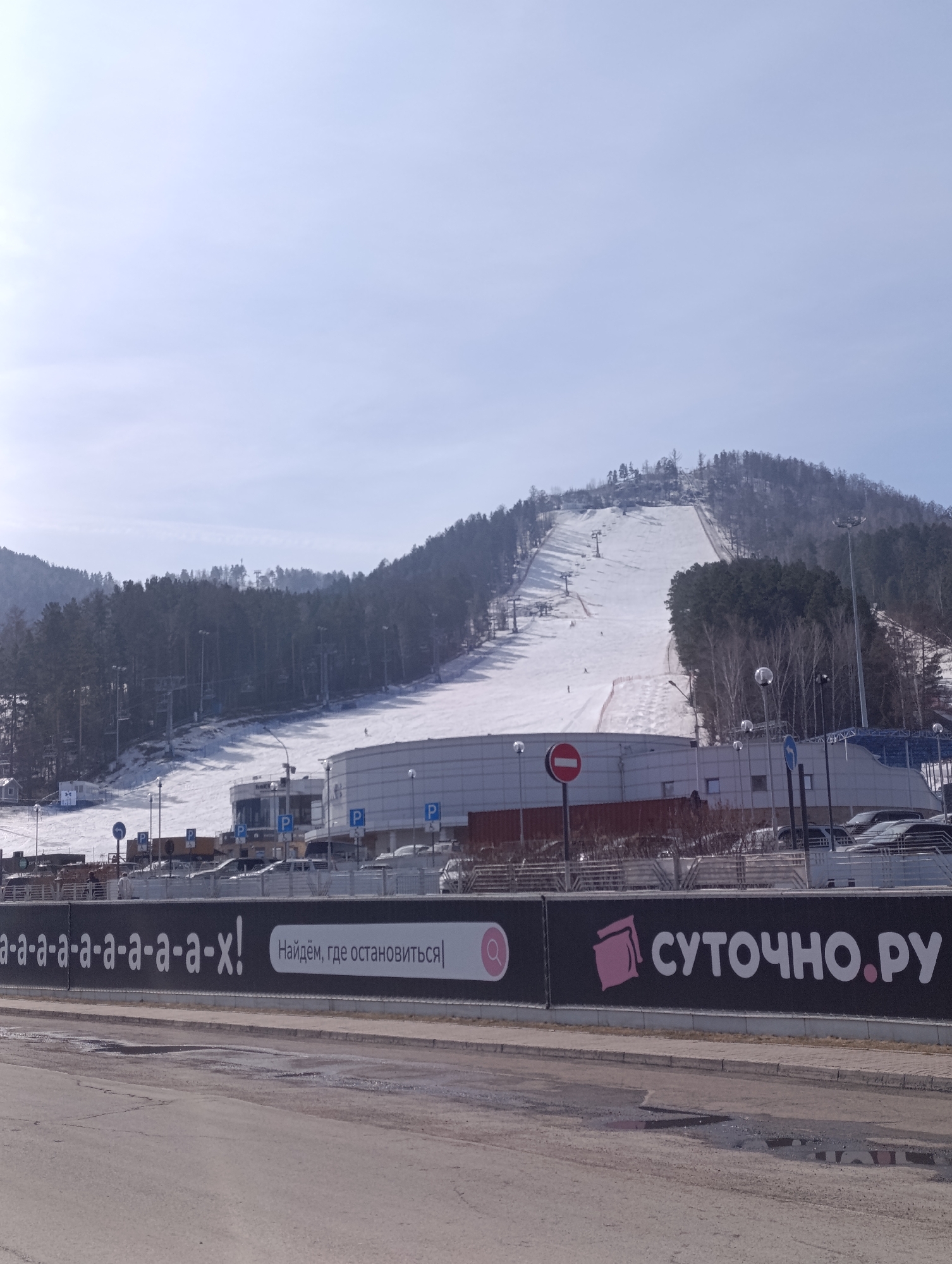 Here is a description of the image:
The image captures an urban scene near a ski resort in Krasnoyarsk, Russia. In the background, a snow-covered mountain with ski slopes and chairlifts is visible. The foreground features an asphalt road with some puddles. A black billboard is situated between the road and the resort, displaying white Cyrillic text on the left, a pink image on the right, and a pink magnifying glass. Behind the billboard, a low-rise building with parking spaces, marked with blue "P" signs, is apparent. The weather appears clear with a bright sky. Several bare trees and lamp posts are also scattered throughout the scene. Some parked cars are visible.