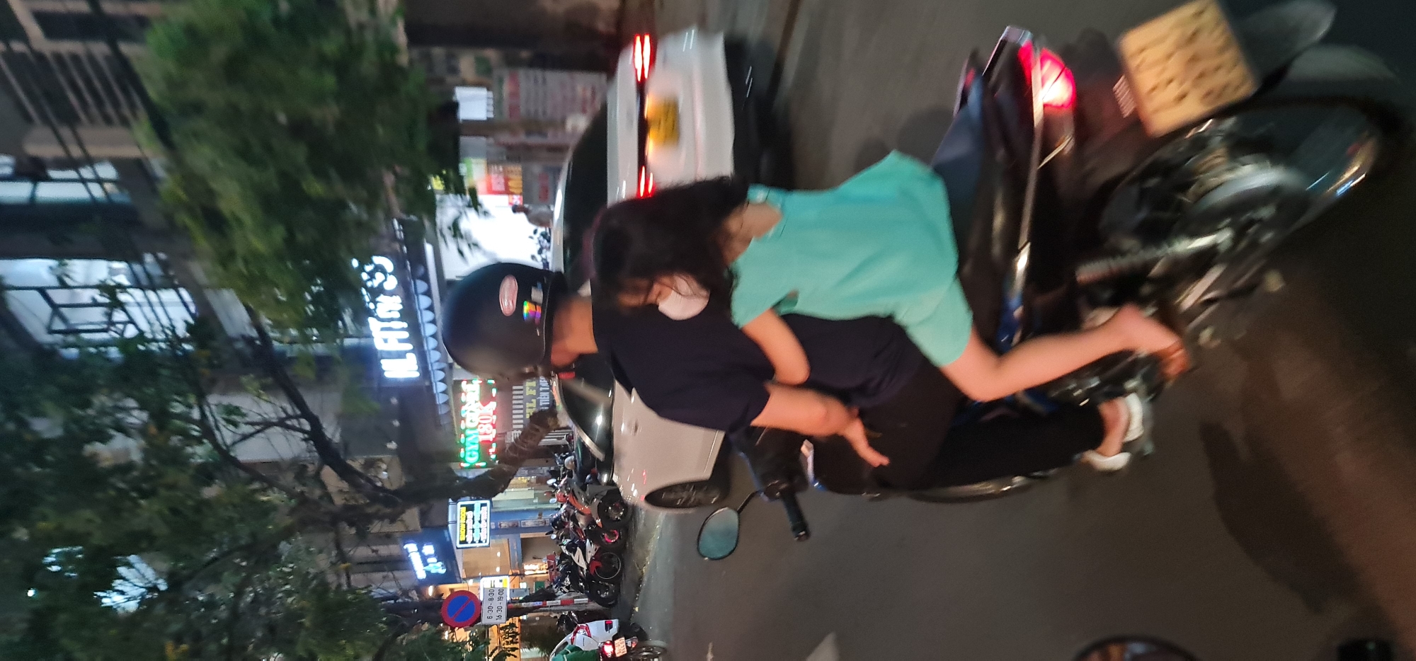 Here's a detailed description of the image:
**People:**
* A person wearing a black helmet is riding a motorcycle.
* A child in a light blue shirt is sitting in front of the rider.
* Both appear to be wearing face masks.
**Subjects:**
* A motorcycle, which is the central mode of transportation.
* A white car is partially visible in the background.
* Several parked motorcycles line the street.
**Objects:**
* The rider wears a black helmet.
* Various street signs are visible. One includes the times 6:30-8:30, and 16:30-19:00.
* A building is visible with the text "IL FAT" printed on it.
**Actions:**
* The rider is operating the motorcycle.
* The child is being transported on the motorcycle.
**Interactions:**
* The child clings to the rider for support.
**Environment:**
* The scene is an urban street.
* It is nighttime.
* The setting is Đà Nẵng, Vietnam, as indicated by the street signage.
* Trees and buildings line the street.
* Streetlights provide illumination.