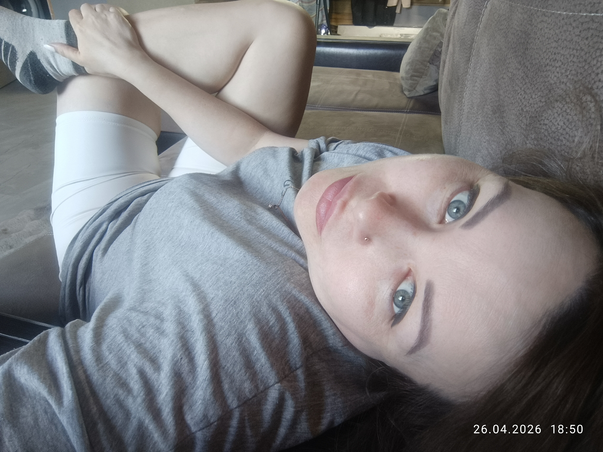 A woman is lying on a brown sofa. She is wearing white shorts, a gray t-shirt, and gray socks with dark stripes. Her right leg is raised and bent at the knee, with her hand holding her foot. She has fair skin, blue eyes, and dark eyebrows. There is a stud piercing on the left side of her nose. The background includes a glimpse of a room with clothes hanging and other furniture. The date and time "26.04.2026 18:50" are visible in the lower right corner. Location is specified as Chaikovsky, Russia.