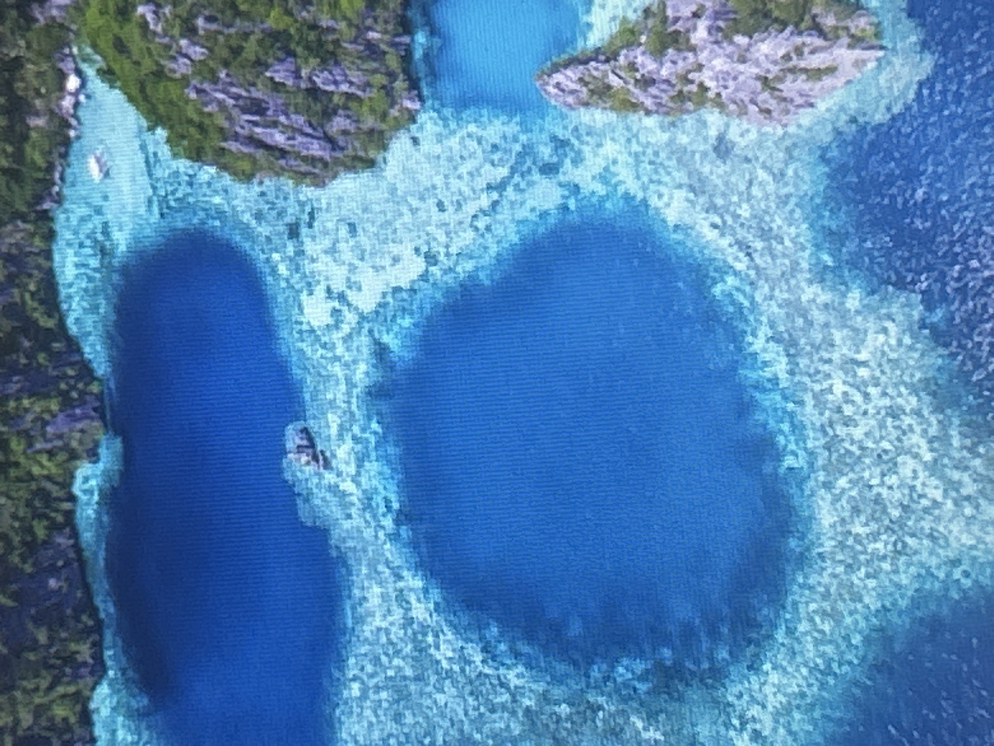This is an aerial view of a tropical lagoon. The water is a vibrant blue and appears clear, with visible coral formations beneath the surface. There are several islands covered in lush green vegetation and rocky outcroppings surrounding the lagoon. In the water, a small white boat is visible in the upper right portion of the image, and a larger vessel, possibly a ferry or tour boat, is positioned in the upper left quadrant. The overall scene depicts a serene and picturesque coastal landscape. There is no indication that this image is located in Sofia, Bulgaria.