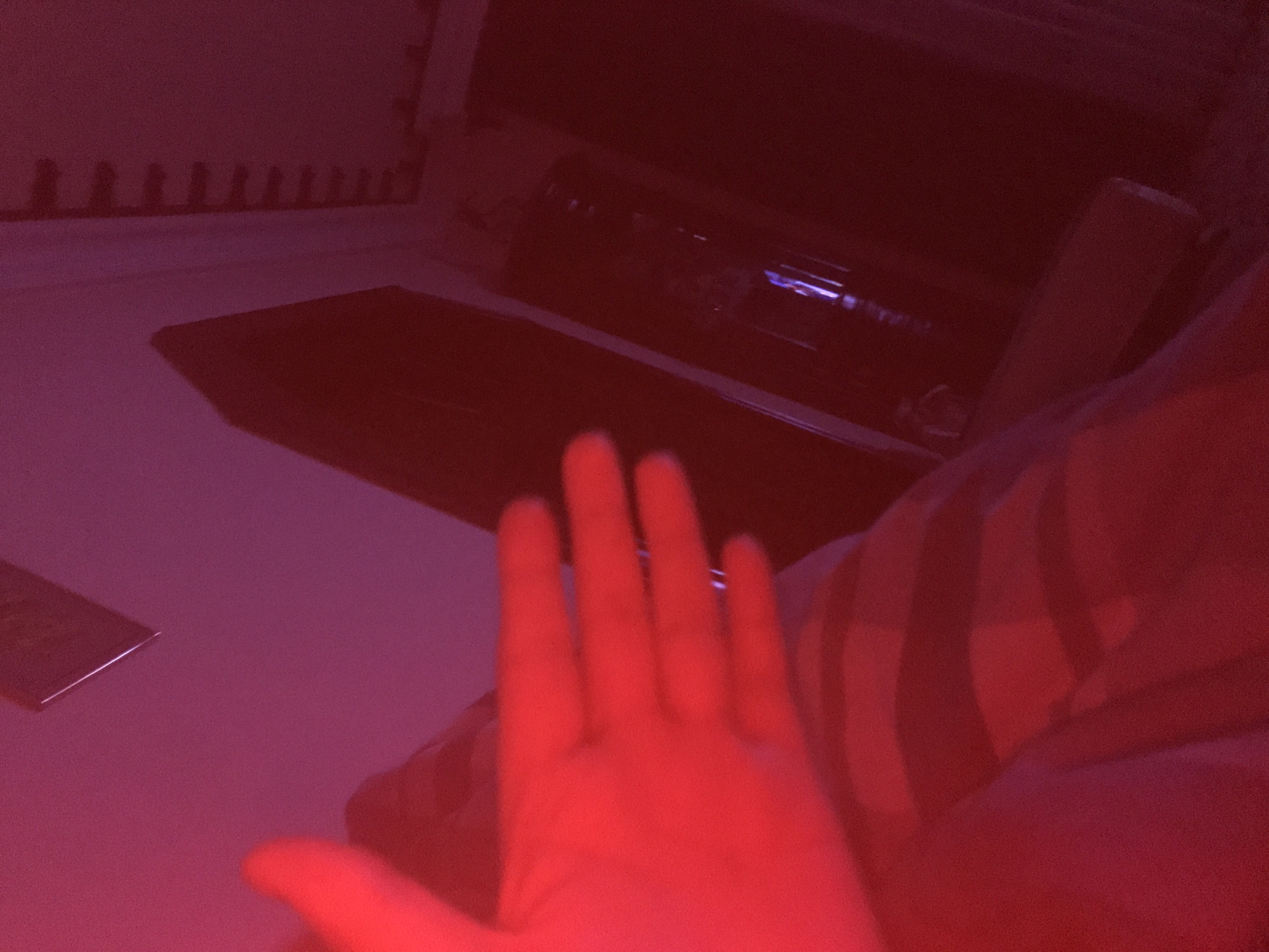 The image shows a dimly lit indoor scene with a strong red cast. A hand is prominently displayed in the foreground. Behind the hand, there appears to be a laptop or similar device on a surface, potentially a desk or table. In the background, indistinct objects are visible, possibly including a window and other furniture. The location of the scene is unknown. There are no clear indicators of it being in Yerevan, Armenia.