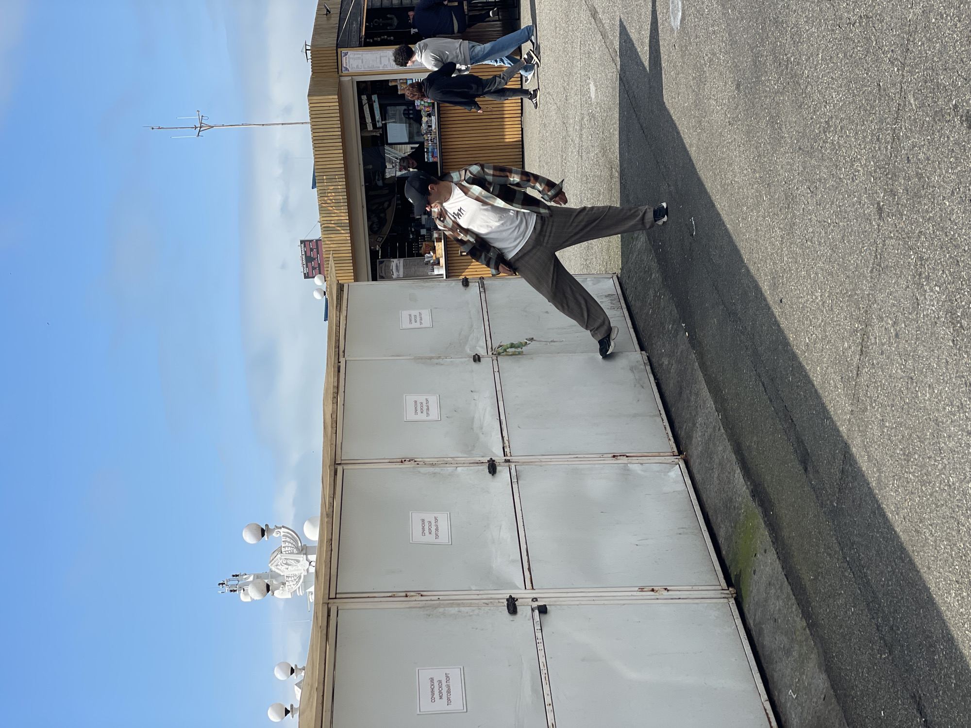 A man stands in front of a booth. The booth is closed and has signs on it that say "SOCHINSKIY MORSKOY TORGOVY PORT." The location is Sochi, Russia. There are other people in the background walking past the booth.