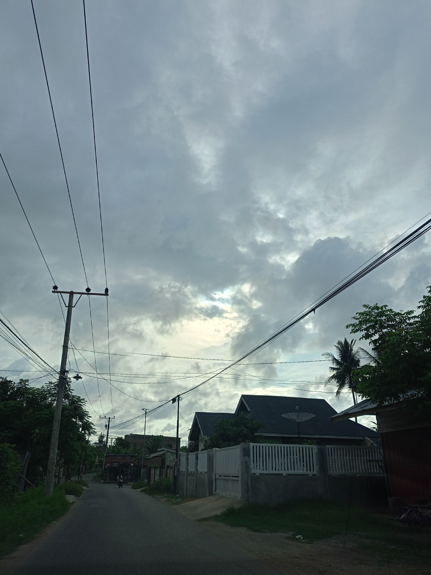 The image depicts a street in Meunasahteungkudigadong, Indonesia, under a cloudy sky. An asphalt road stretches into the distance. The street is lined with residential buildings, most enclosed by fences. Overhead, a network of electrical wires is strung between utility poles. A single motorcyclist is visible on the road in the distance. The scene appears quiet and overcast. A palm tree is visible among other vegetation.